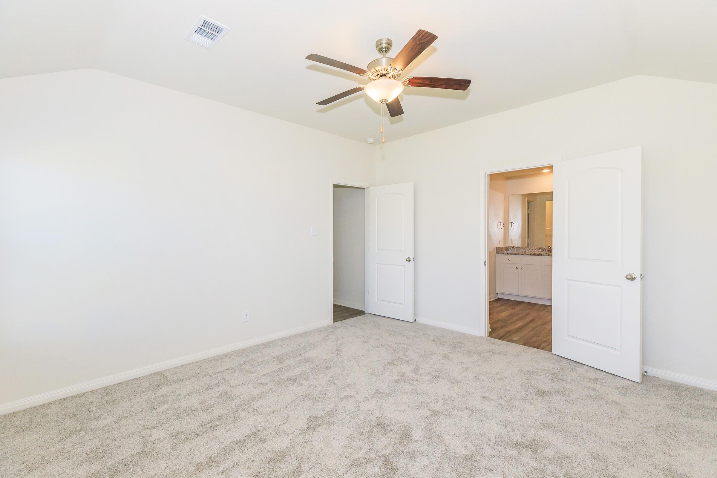 A bright, empty bedroom featuring light beige carpet, a ceiling fan with wooden blades, and two white doors leading to adjoining spaces. The walls are painted in a light color, enhancing the spacious feel of the room. The room is well-lit with natural light from a nearby window.