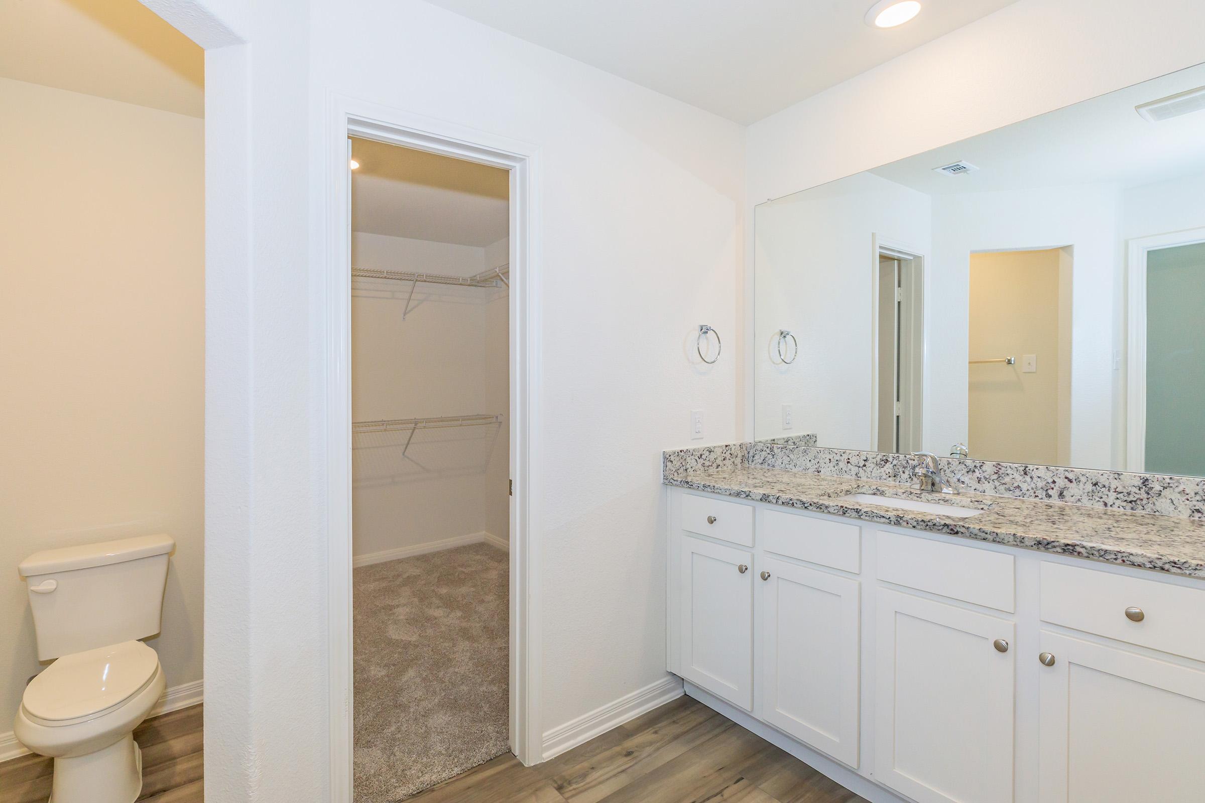 A modern bathroom featuring a double sink vanity with a granite countertop, a bright mirror, and a closet space. There's a doorway leading to a walk-in closet and a separate toilet area. The flooring is light-colored, complementing the clean, minimalistic design of the space.