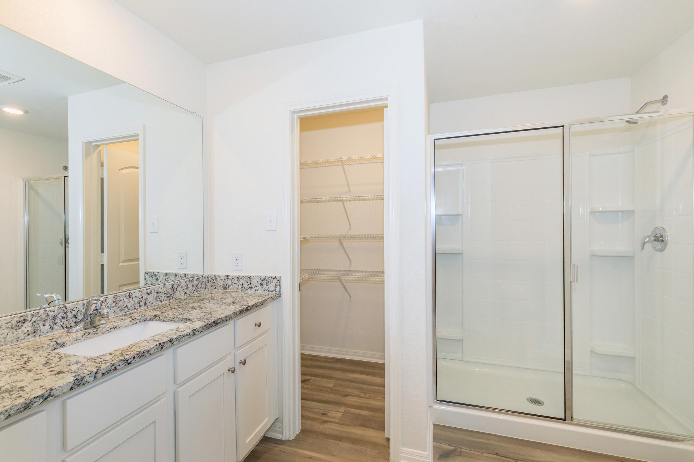 Bright and modern bathroom featuring a granite-topped vanity with a sink and ample storage, a large walk-in closet with shelving, and a glass-enclosed shower. The walls are painted white, and the flooring is a light wood finish, creating a clean and inviting atmosphere.