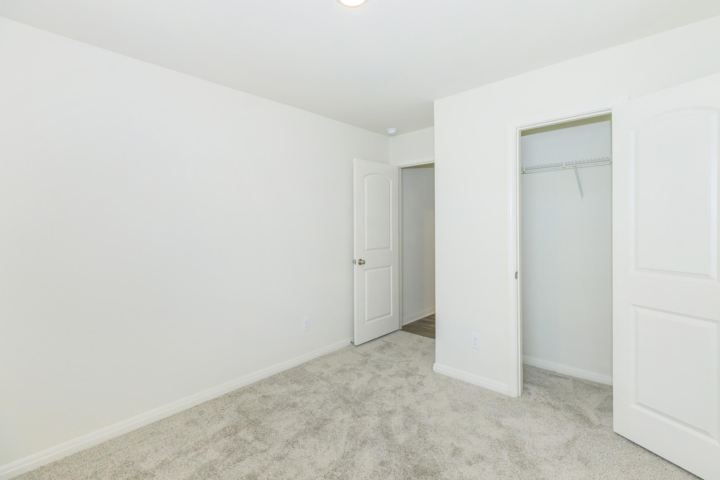 A light-colored empty room with beige carpet flooring, featuring two doors: one leading to a closet and the other to an adjacent space. The walls are painted white, giving the room a clean and bright appearance. A ceiling light provides additional illumination.