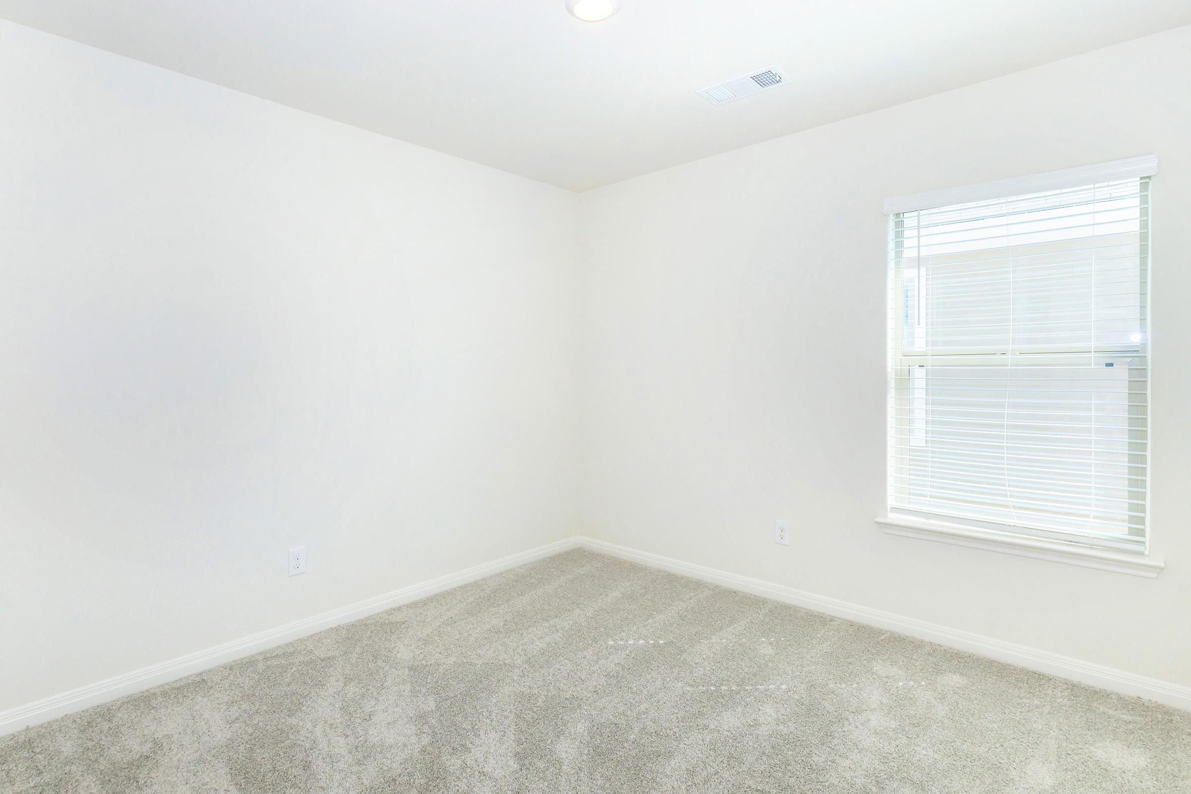 A vacant room featuring light beige walls, a window with white blinds allowing natural light, and soft gray carpet. The space is simple and uncluttered, providing a blank canvas for personalization.