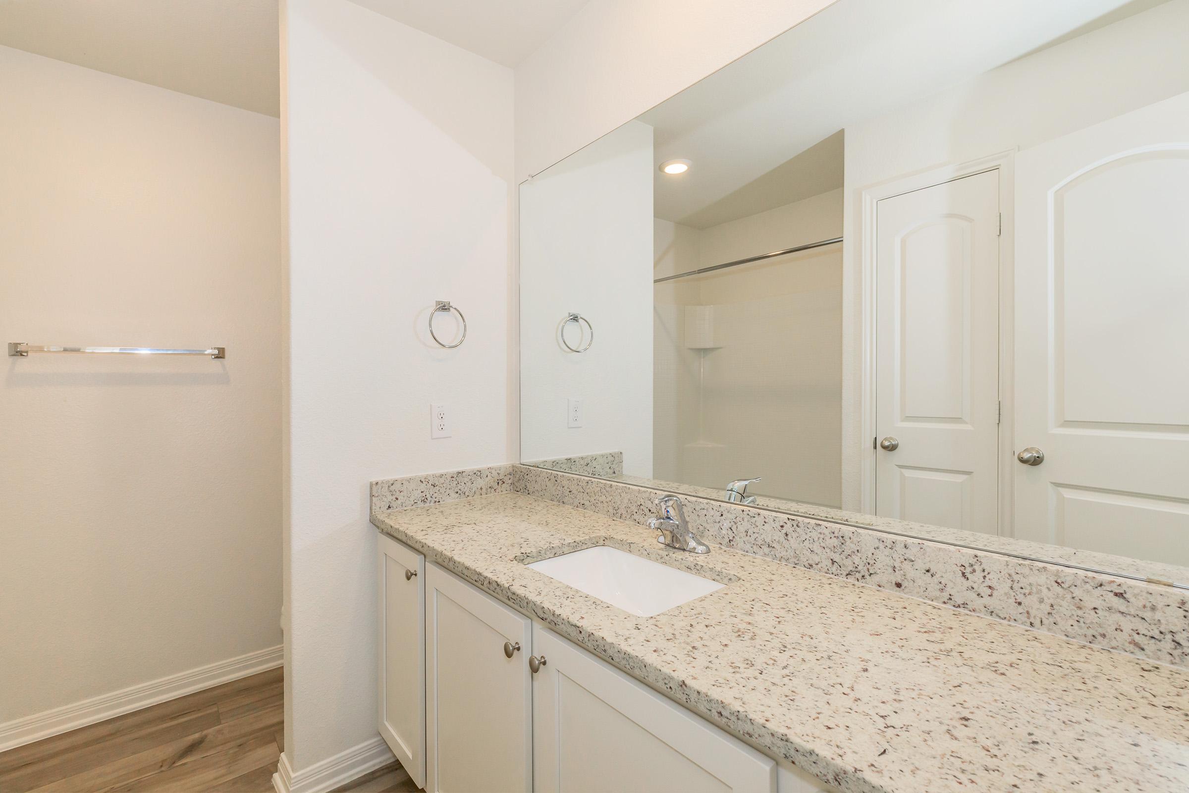 A modern bathroom featuring a light-colored countertop with a sink, silver faucets, and a large mirror. The walls are painted white, with a towel bar on one side. The flooring is wood-like, and there is a shower area visible in the background. The overall design is clean and contemporary.
