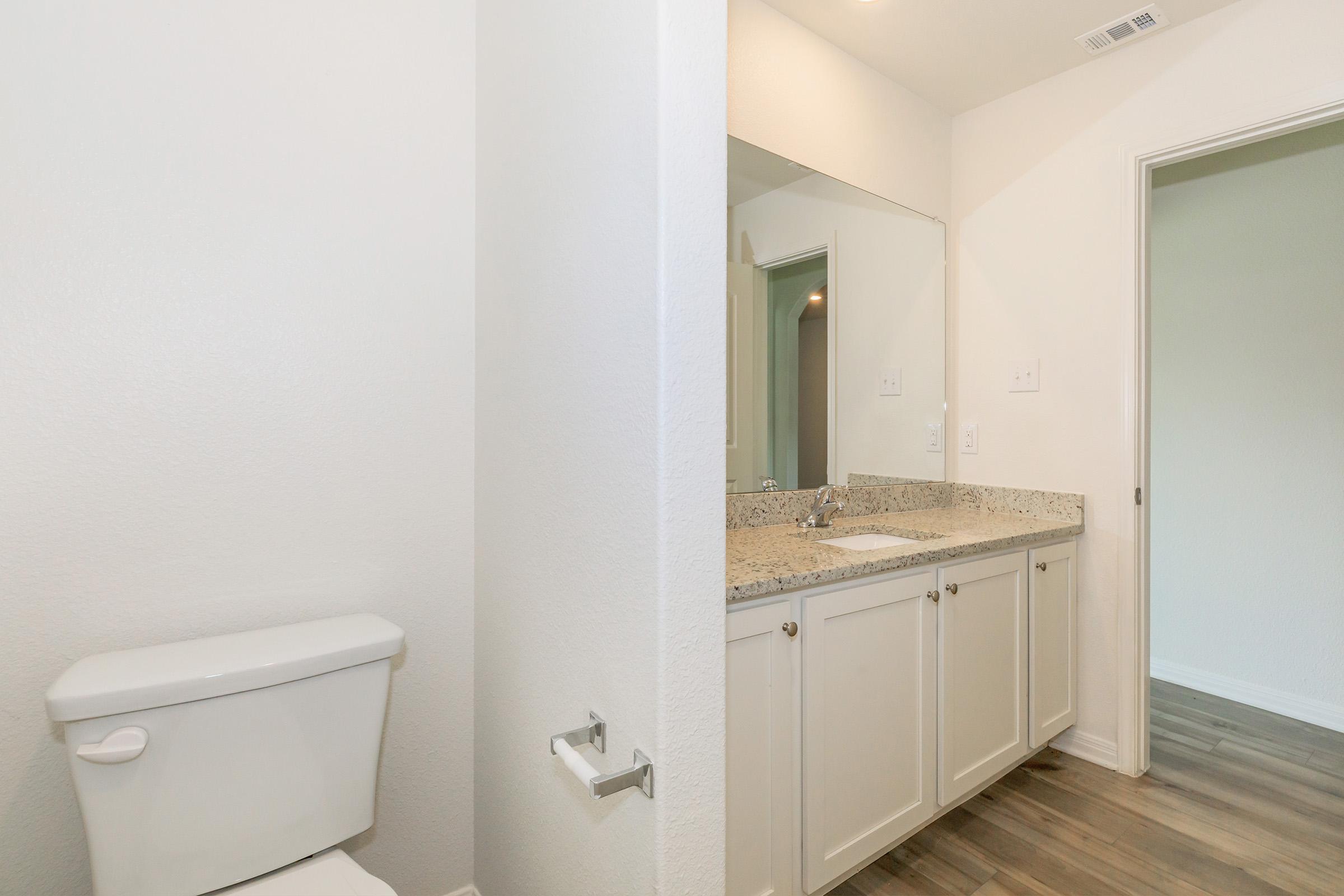 A clean and modern bathroom featuring a white toilet, a light-colored granite countertop with a sink, and a large mirror. The walls are painted in a soft color, and the floor has wooden-looking tiles. There is a doorway on the right leading to another room.