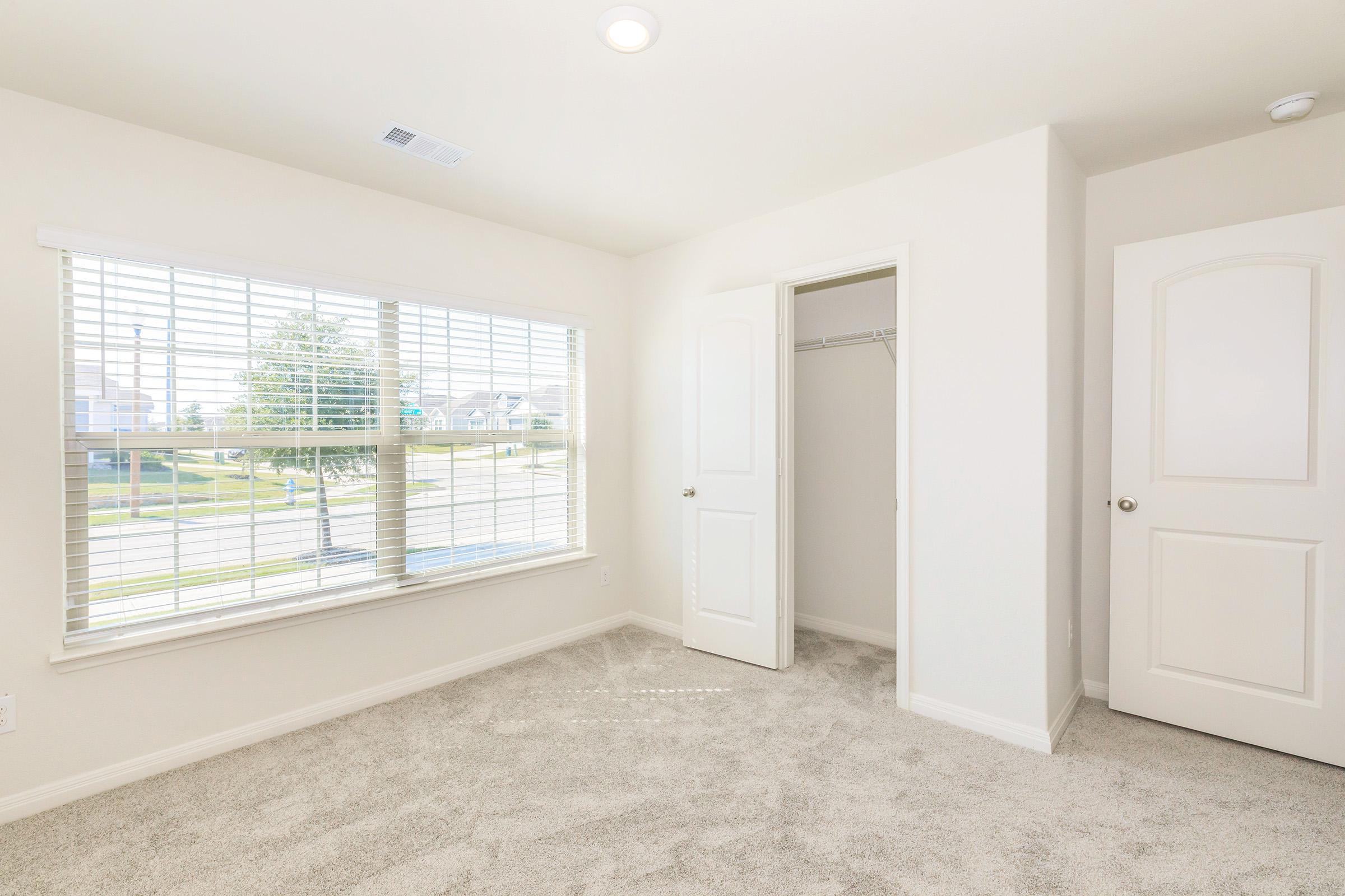A bright, empty bedroom featuring light-colored walls, soft carpeting, and large windows with blinds. There is a closet with double doors on one side and the room is well-lit by natural light. The view outside includes greenery and houses in the distance.