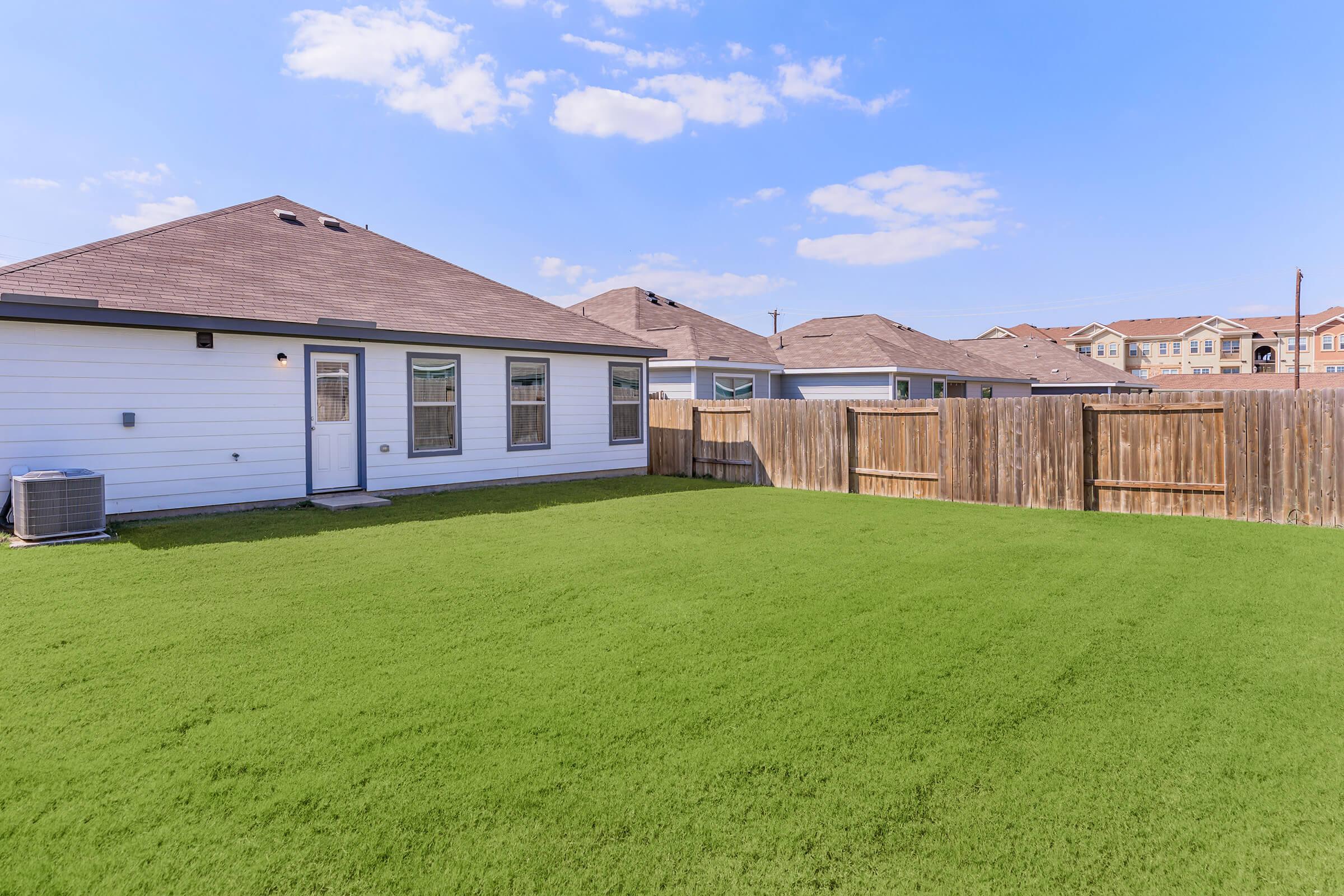 A spacious backyard with freshly cut green grass, surrounded by a wooden fence. In the background, there are several residential houses with rooftops against a clear blue sky. A door leads from the house to the yard, and an air conditioning unit is visible on the side of the house.