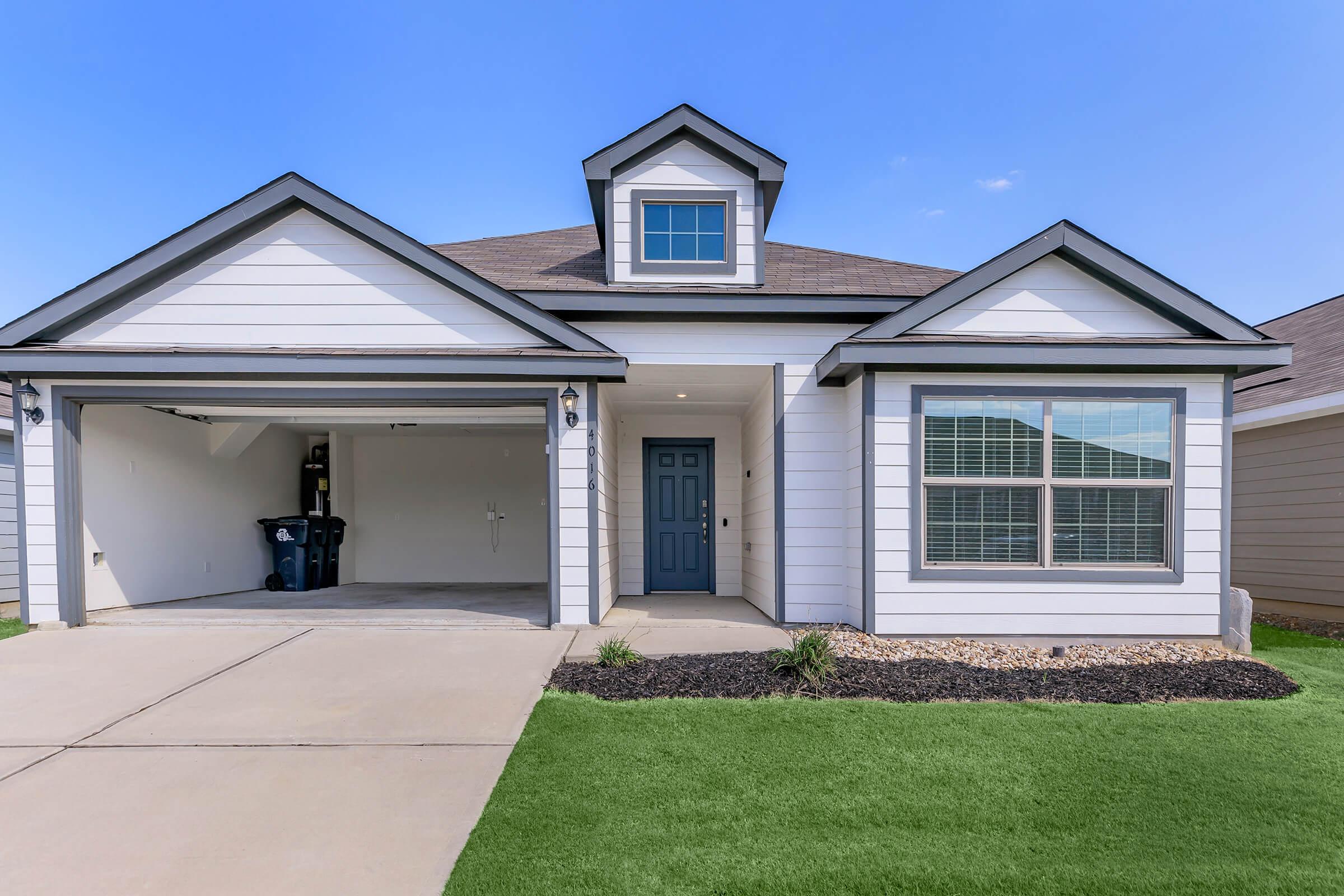 A modern single-story house featuring a light-colored exterior, a peaked roof, and a garage. The front yard has green grass, landscape rocks, and minimal landscaping. A blue door serves as the main entrance, and there are large windows on either side of the door. The driveway is paved, leading to the garage.