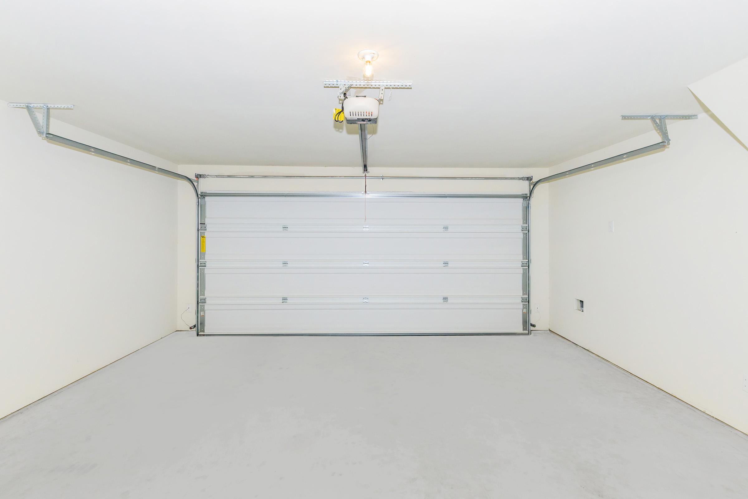 A spacious, empty garage with a white garage door and a ceiling-mounted light fixture. The walls are painted in a neutral color, and the floor is smooth and clean. There are no vehicles or items inside, creating an open and uncluttered space.