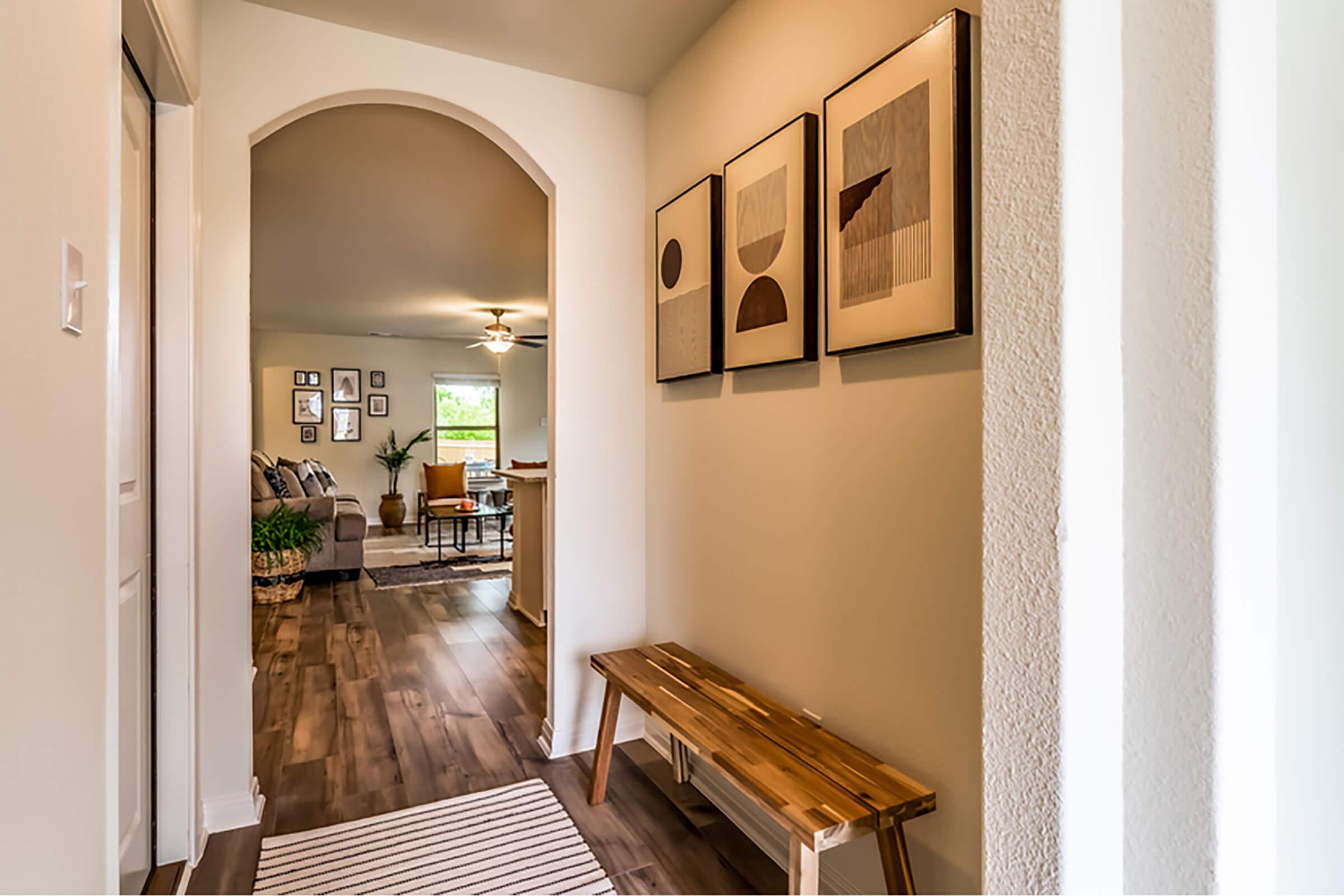 A well-lit hallway featuring a wooden bench and a striped rug, leading towards a living room. The walls are adorned with framed artwork in neutral tones, and a gentle archway transitions to the spacious living area, which includes a cozy couch and plants.