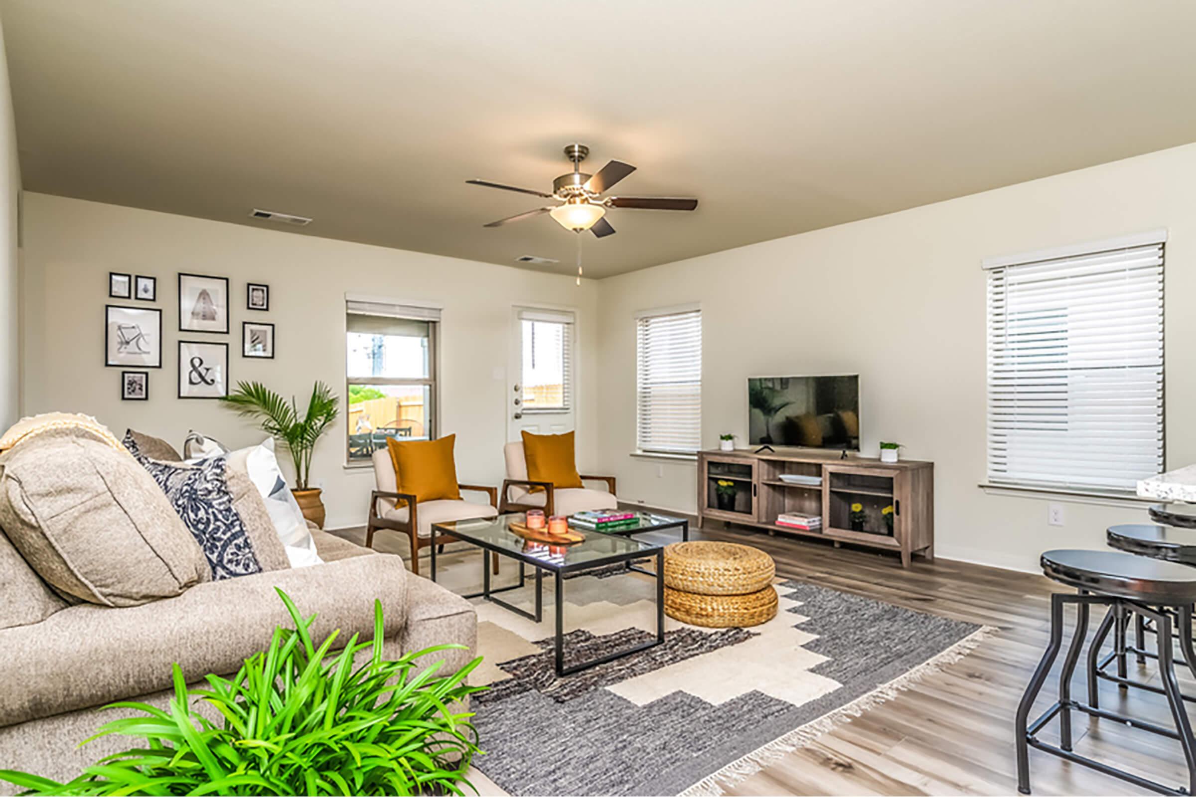 Cozy living room featuring a gray sofa with decorative pillows, a wooden coffee table, and a plant. The space has light walls, wooden flooring, and large windows with blinds. A television is set on a modern console, and artwork is displayed on the walls. Two yellow armchairs add a pop of color to the room.