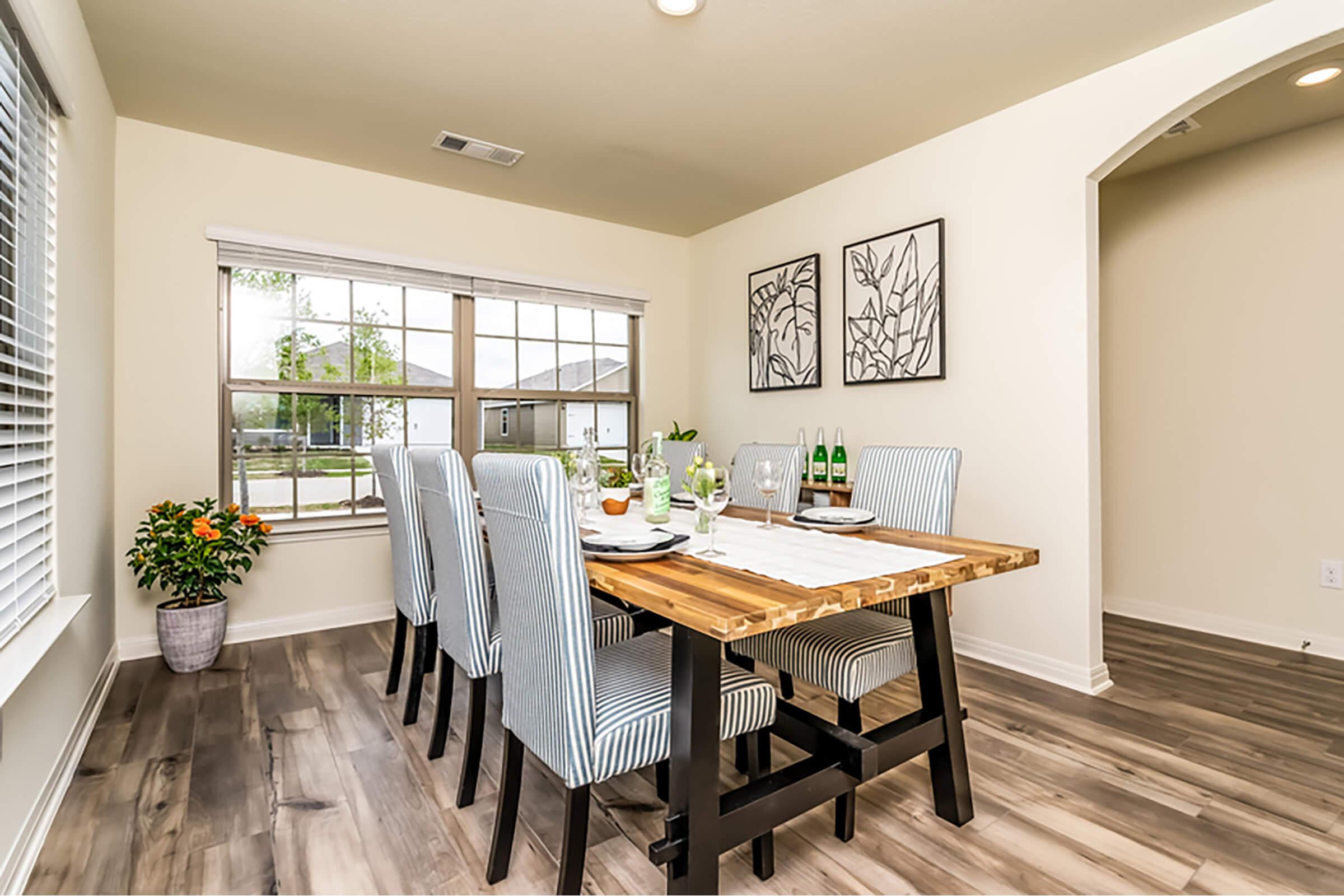 A bright dining area featuring a wooden table set for a meal with plates, glasses, and a vase of flowers. Six upholstered chairs surround the table, and large windows let in natural light. Decorative plant and framed artwork adorn the walls, creating a welcoming atmosphere.