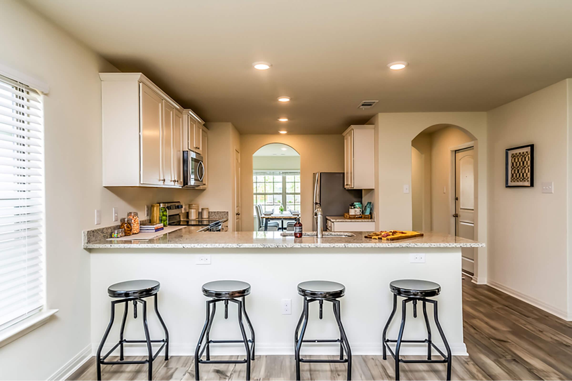 Modern kitchen featuring a bar area with four black stools, white cabinetry, and a granite countertop. The space includes stainless steel appliances and an open layout leading to a bright dining area with large windows. Neutral color palette with ample lighting.