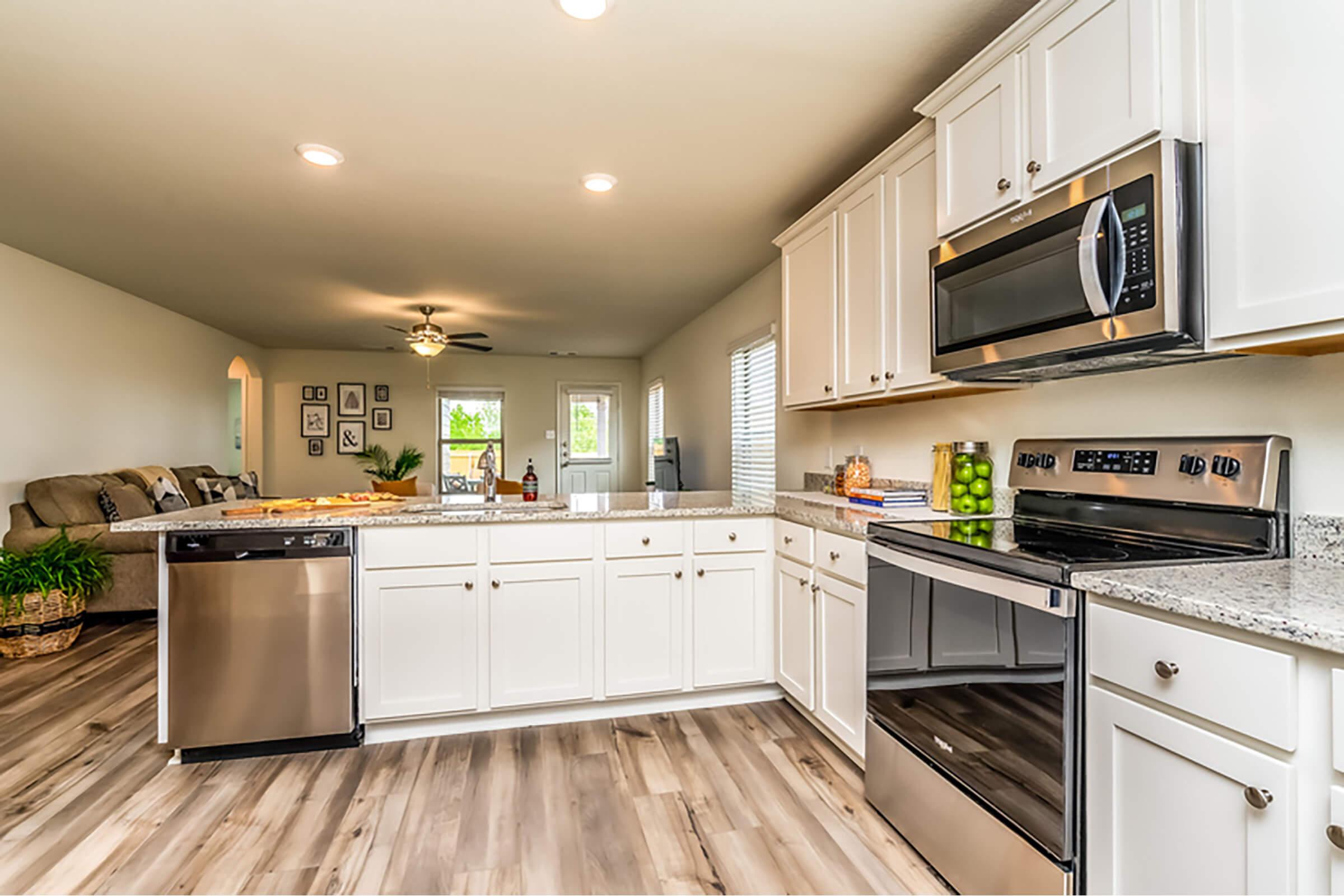 Modern kitchen featuring white cabinetry, granite countertops, a stainless steel microwave, and a dishwasher. The space includes wood-style flooring and has open access to a cozy living area in the background, filled with natural light and a comfortable seating arrangement.