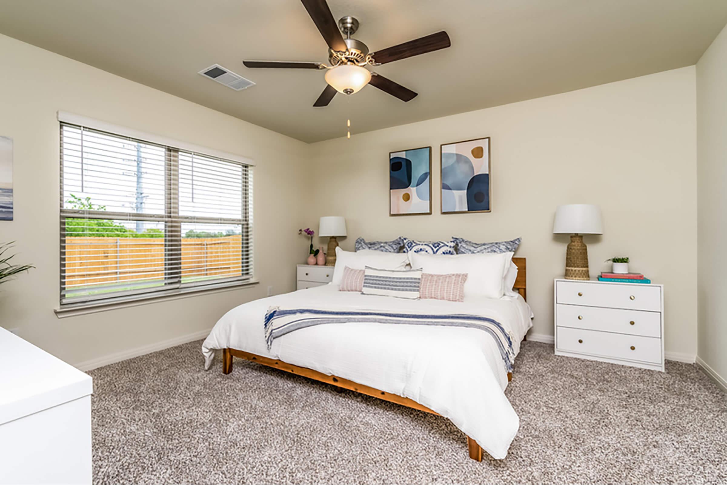 Cozy bedroom featuring a large bed with white bedding and decorative pillows, a stylish ceiling fan, and two bedside tables with lamps. The walls are a soft cream color, and there are two framed artworks above the bed. Large windows provide natural light, with a view of a fenced outdoor area.