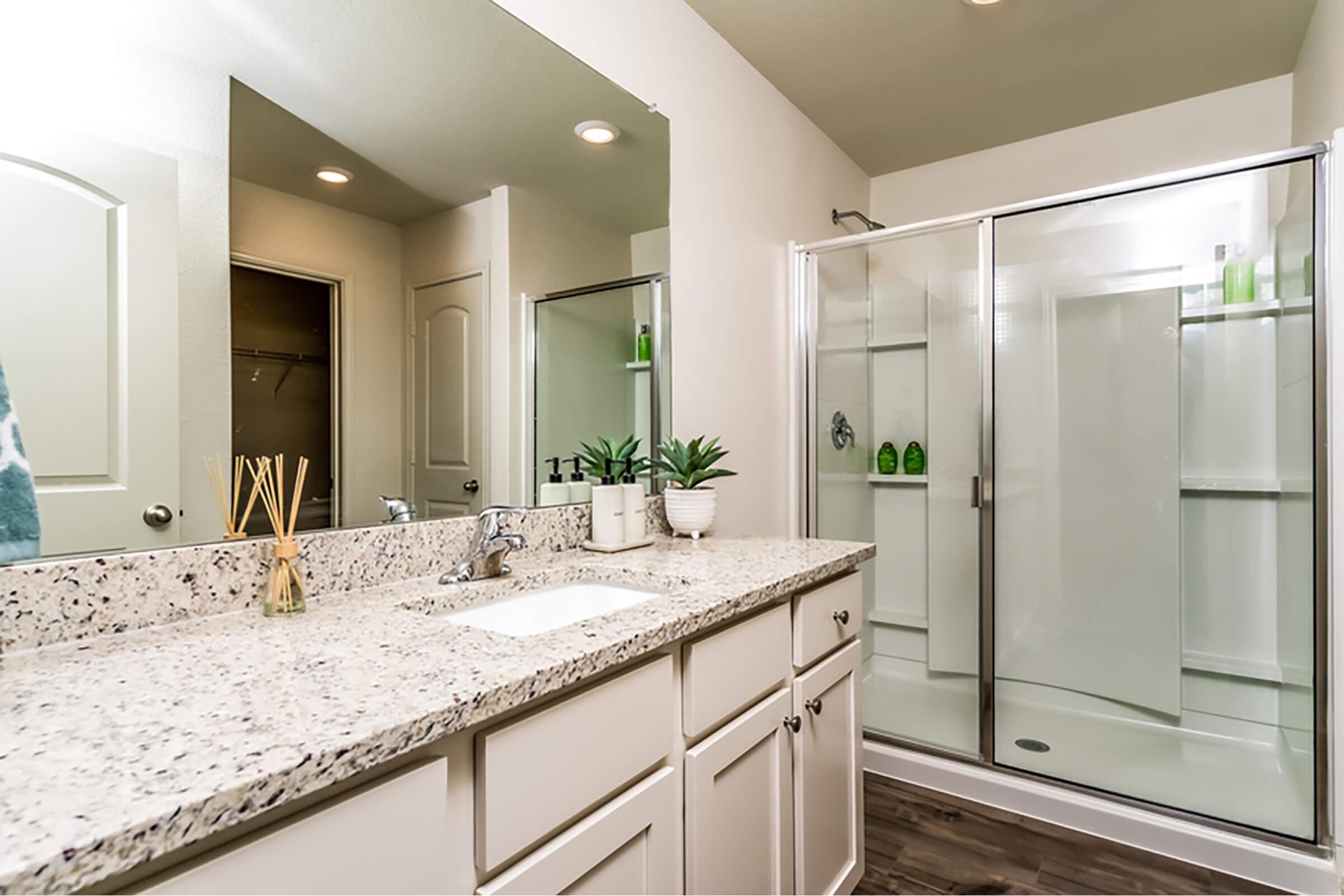 Modern bathroom featuring a double-sink vanity with speckled countertop, a large mirror, and stylish decor. A glass shower enclosure is visible, alongside storage shelves. Soft lighting creates a welcoming atmosphere, complemented by potted plants for a touch of greenery.