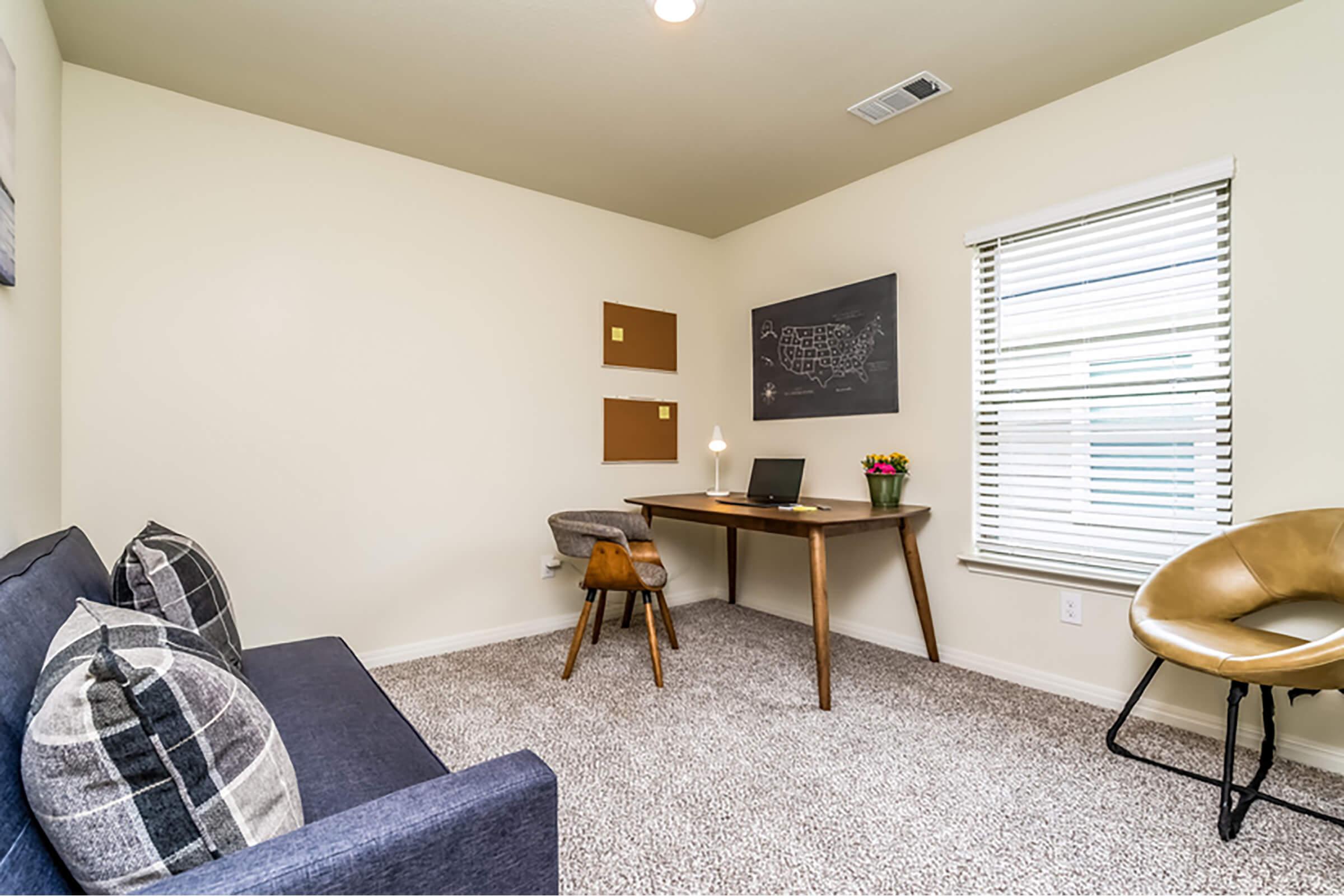 A small home office featuring a dark wood desk with a laptop, a chair, and a lamp. There is a cozy sofa with pillows, a chalkboard on the wall, and two wall-mounted boards. A window with blinds allows natural light to enter, and a small potted plant adds a touch of greenery to the space.