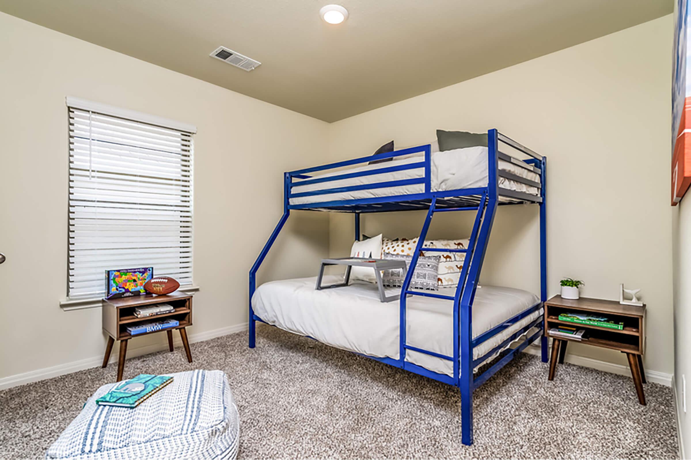 A cozy bedroom featuring a blue metal bunk bed with a top and bottom sleeping area, light-colored walls, and carpet flooring. There are two small wooden nightstands beside the bed, one with a decorative plant, and a window with white blinds allowing natural light. A colorful wall art piece is visible.