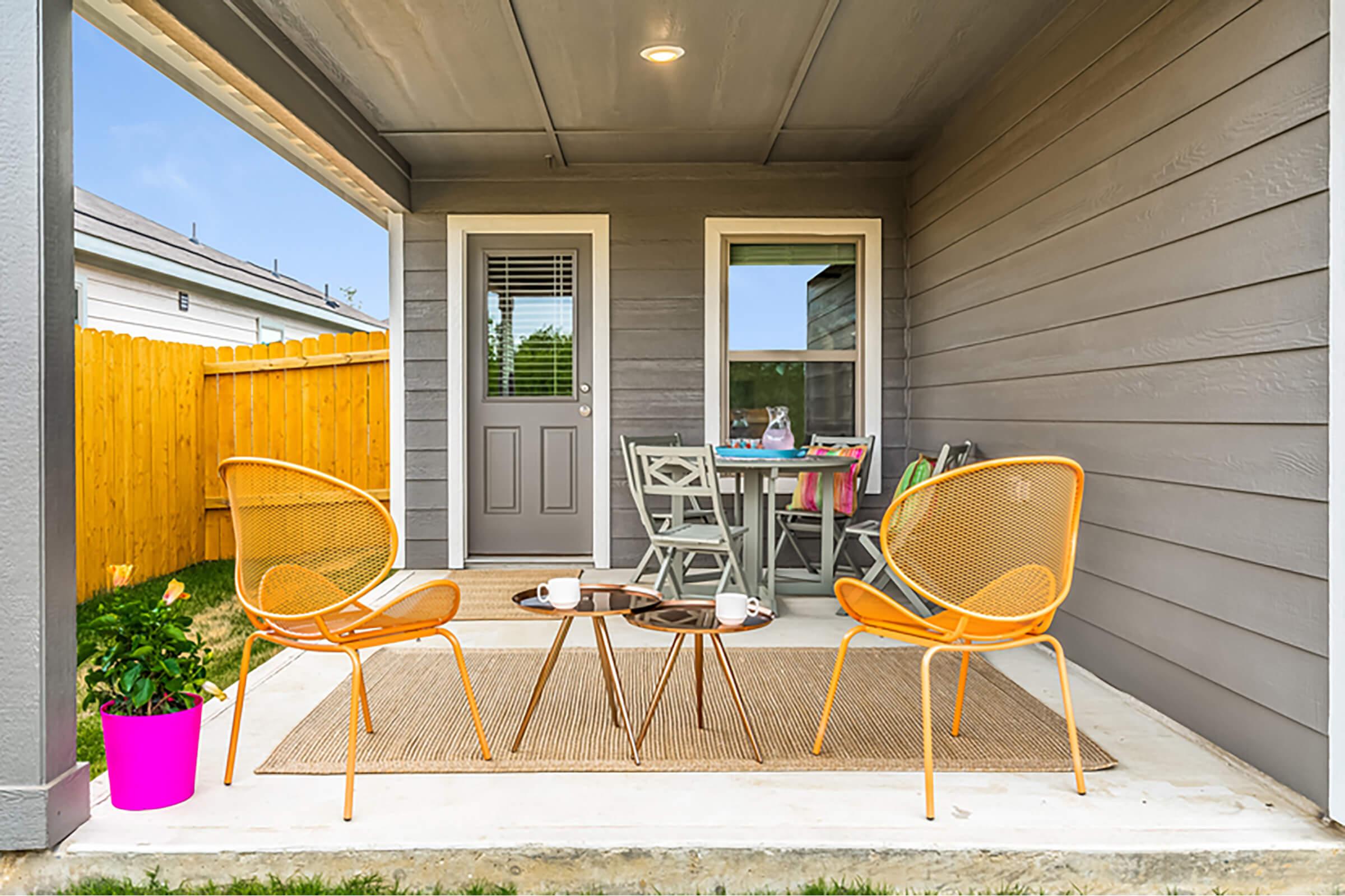 A cozy porch with two stylish orange chairs and a small round table. A pink planter with greenery sits nearby. In the background, there is a wooden fence, a door, and a dining table with chairs. The space is well-lit, creating a welcoming outdoor seating area.