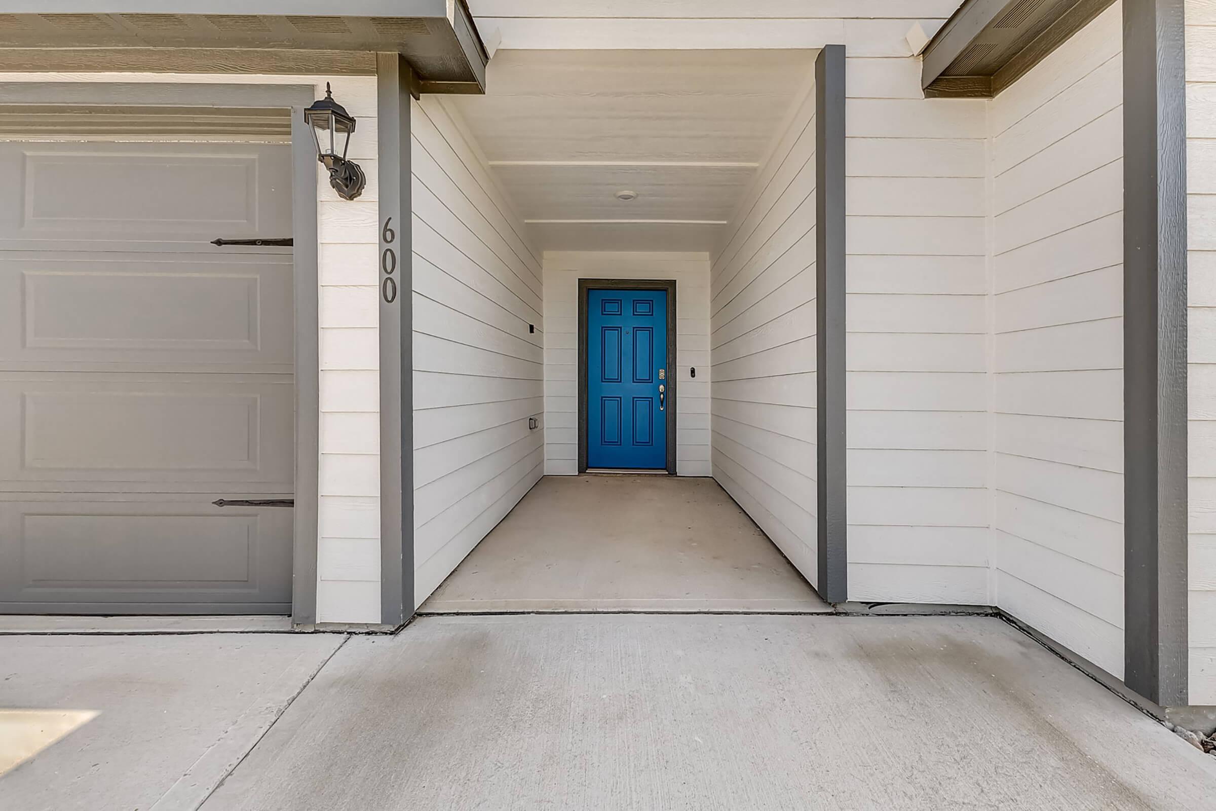 A house entrance featuring a light gray garage door and a bright blue front door. The entryway has white siding and a concrete walkway leading up to the door, flanked by vertical gray columns. There is a wall-mounted light fixture beside the door.