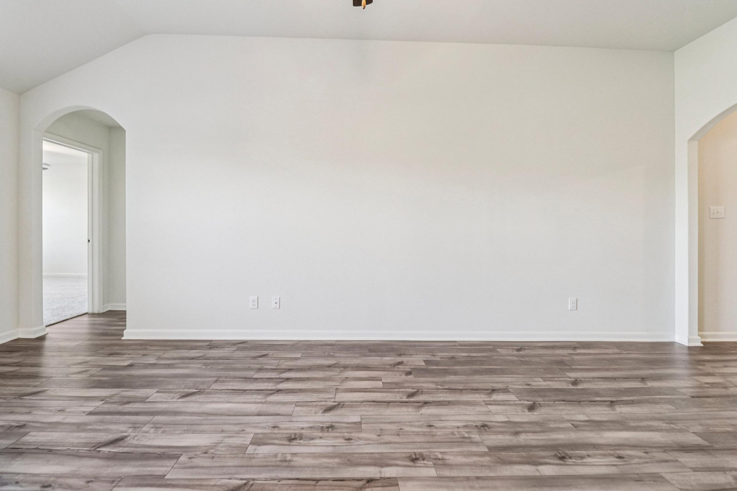 A spacious, empty room with light-colored walls and a wood laminate floor. Two archways lead to adjacent rooms, and the ceiling features a ceiling fan. The overall ambiance is bright and airy, ideal for interior decoration or furniture arrangement.