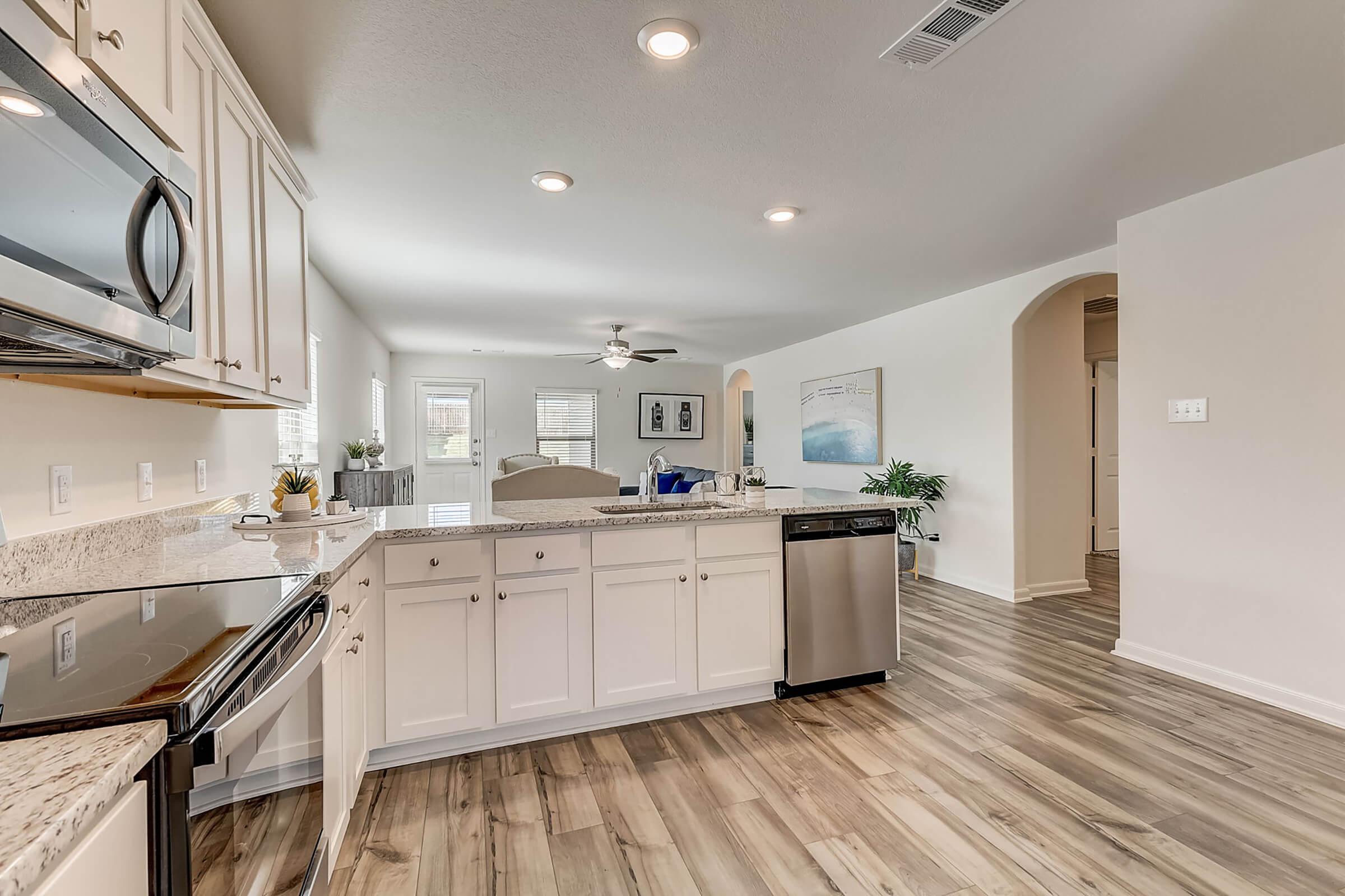 A modern kitchen featuring white cabinetry, granite countertops, and stainless steel appliances. The space includes a large sink, an island with seating, and is open to a bright living area with natural light. Light wood flooring runs throughout the area, enhancing the contemporary design.