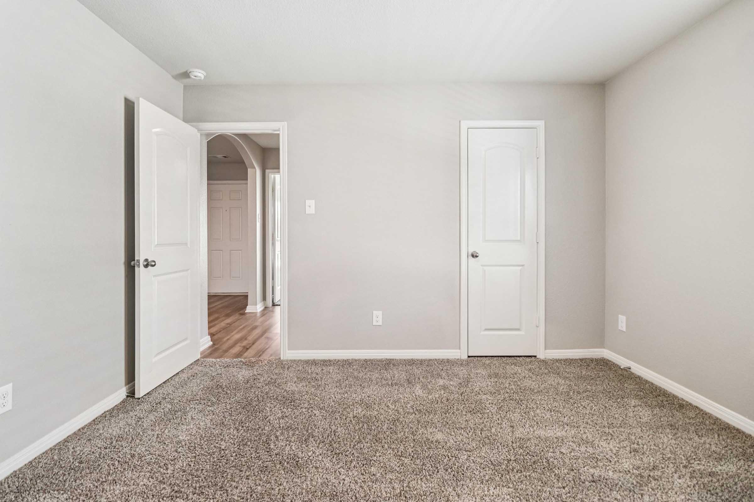 A spacious, well-lit room with light gray walls and plush carpet. Two doors are visible: one leading to another room and another closet door. The room has no furniture, emphasizing the open space and clean lines, making it ready for personal customization.