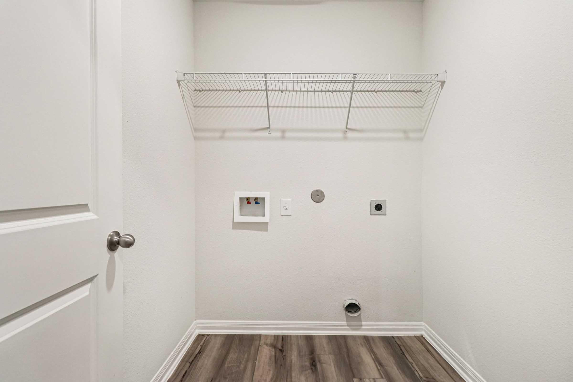 A small, empty laundry room featuring a white wall and a wire shelf mounted on the wall. There are two electrical outlets, and a vent on the floor, with light-colored wooden flooring. The room is devoid of any appliances or items, giving it a clean and minimalistic appearance.