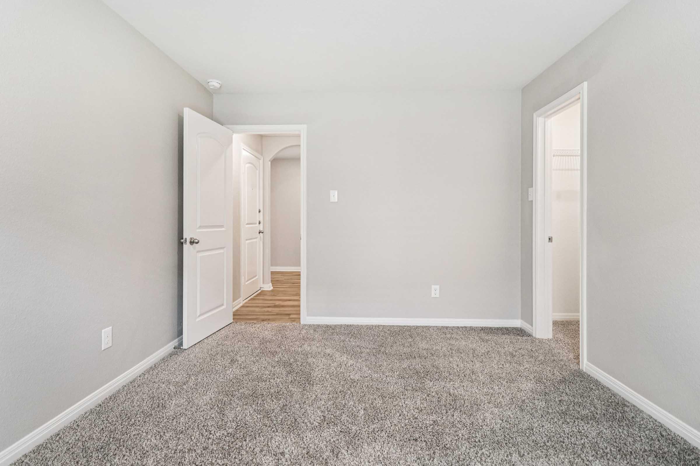 A spacious, empty room featuring light gray walls and carpeted flooring. Two doorways lead to adjacent areas—a door on the left and an open archway on the right. Natural light illuminates the space, creating a clean and inviting atmosphere. Perfect for personalization or as a new living area.