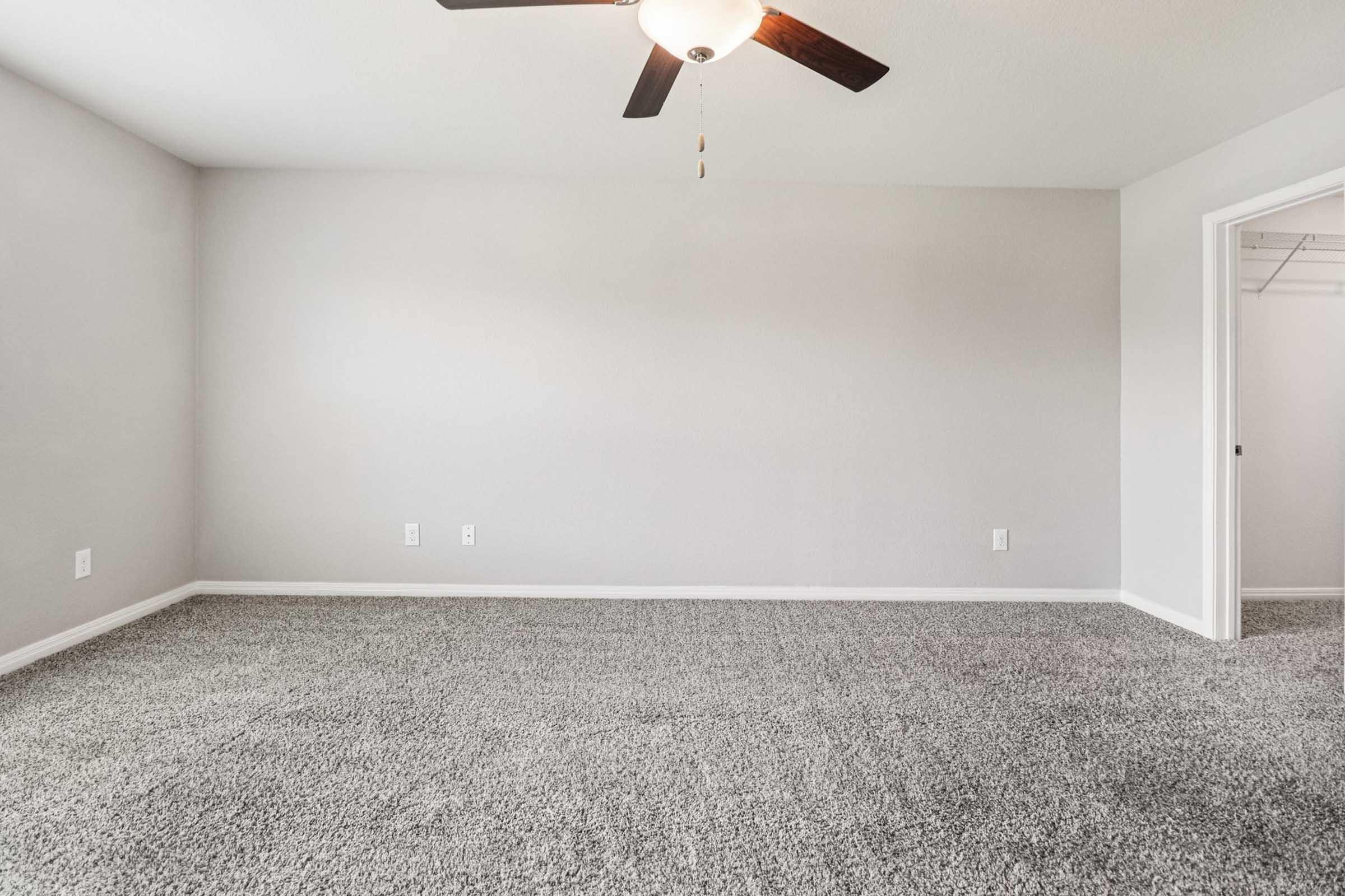 A spacious, empty room with light gray walls and plush, light gray carpet. A ceiling fan hangs from the ceiling, providing a modern touch. To the right, an open door leads to a closet. The overall atmosphere is bright and inviting, ideal for personalizing with furnishings.
