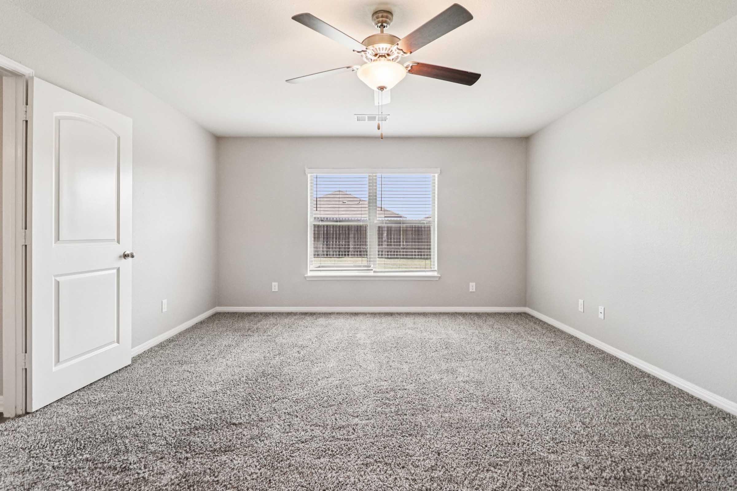 A spacious, empty bedroom with light gray walls and plush carpet. A ceiling fan is mounted above, and a large window allows natural light to enter, showcasing an outdoor view. The room features a door on the left, emphasizing its open and airy feel.