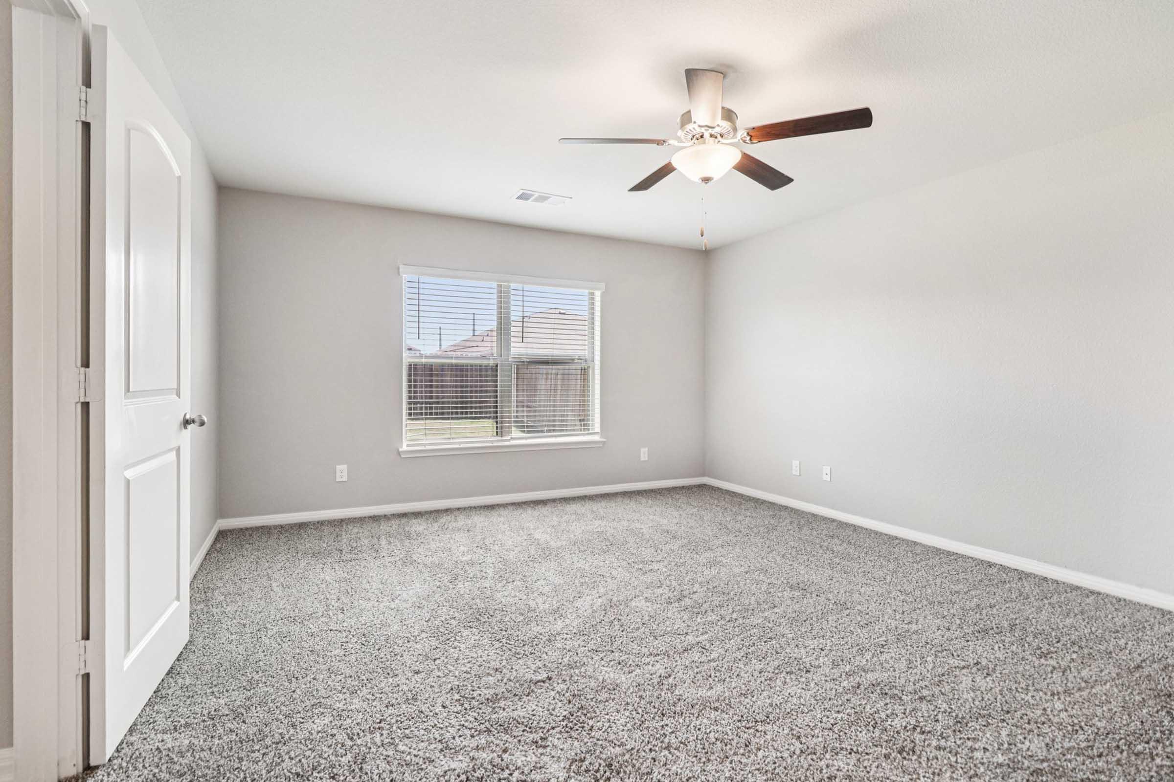 A spacious, empty room with light gray walls and a ceiling fan. The floor is covered in plush gray carpet. A large window lets in natural light, showing a view of the outdoors. A door on the left leads to another room or area. The overall atmosphere is bright and inviting.