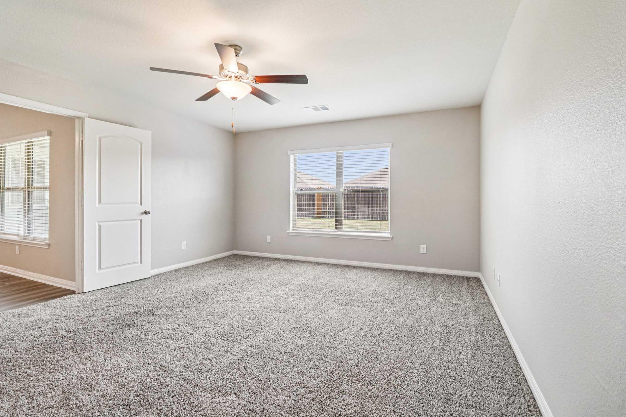 A spacious, well-lit room with light gray walls and plush carpet. It features a ceiling fan and a large window with blinds, offering a view of the outdoors. An open door leads to another area, enhancing the airy feel of the space. Perfect for a living or bedroom setting.