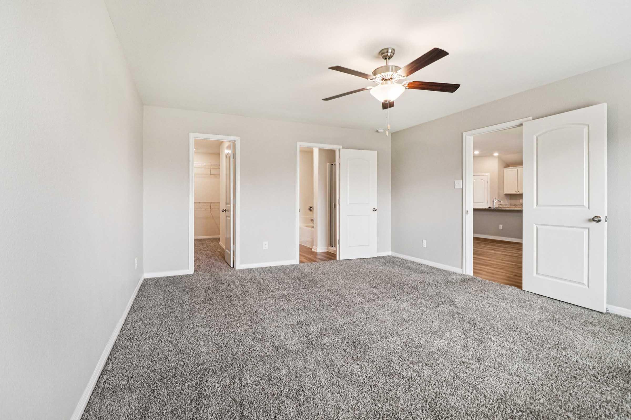 A spacious, carpeted room with a ceiling fan, featuring multiple doorways leading to adjacent areas. The walls are painted in a light color, providing a bright and airy feel. Natural light enhances the inviting atmosphere, making it an ideal living space.