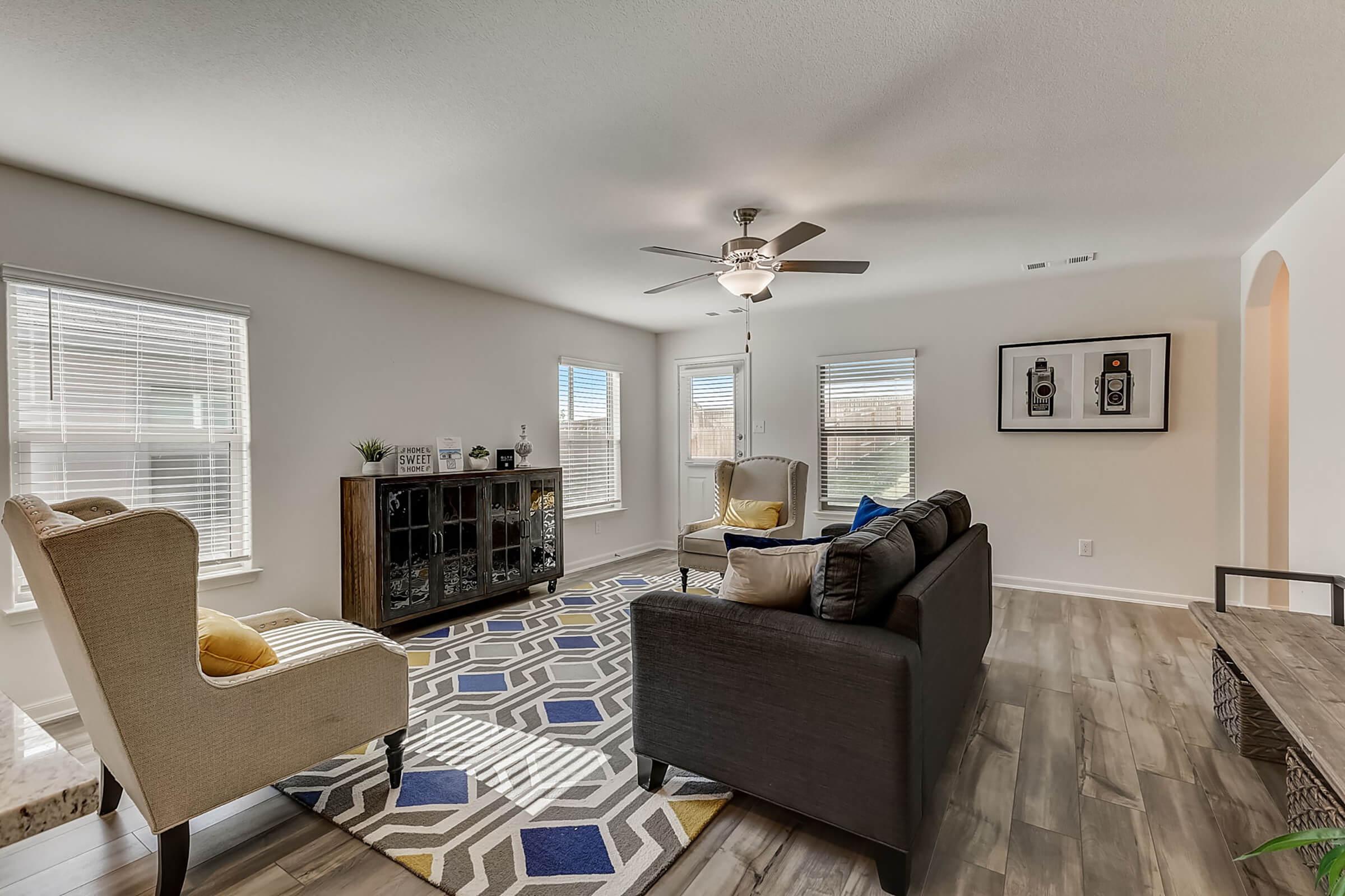 A bright and spacious living room featuring a gray sofa, two armchairs, and a decorative area rug. The room has natural light coming through several windows, and a modern ceiling fan. A stylish cabinet displays decorative items, enhancing the inviting atmosphere.