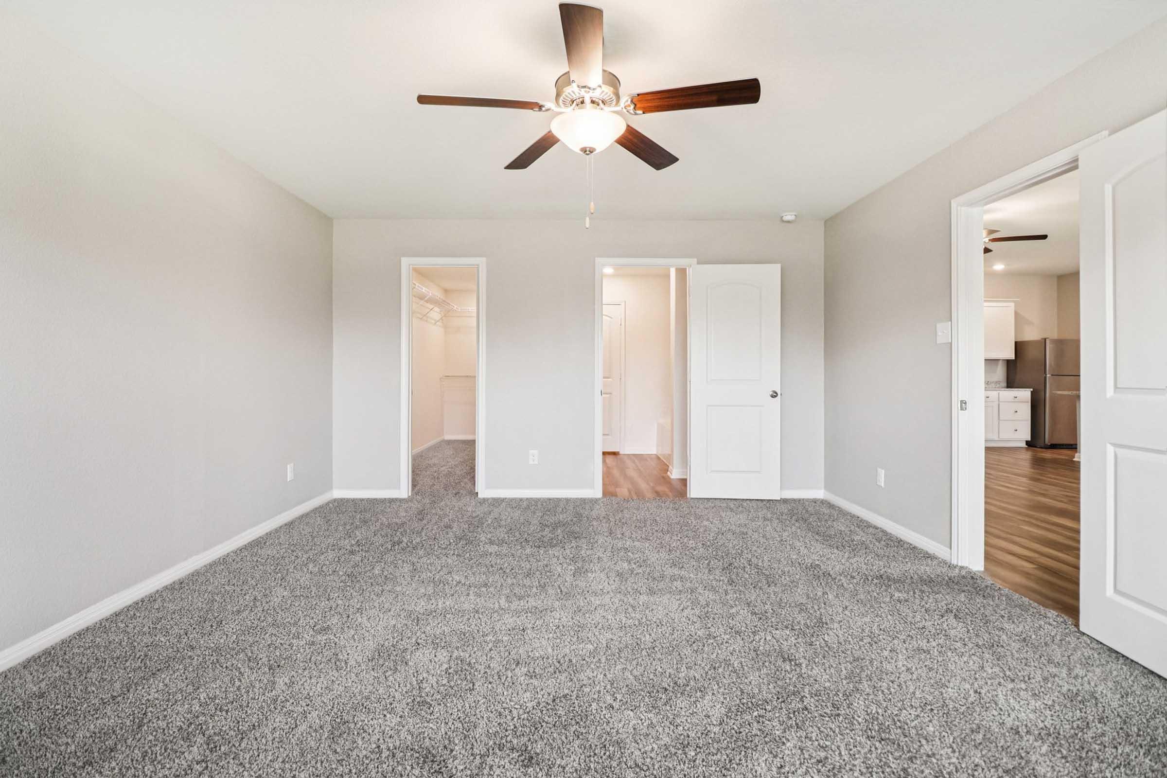 Spacious, carpeted room with a ceiling fan, featuring two open doorways leading to closets and a kitchen area in the background. The walls are painted a light color, creating a bright and airy atmosphere.