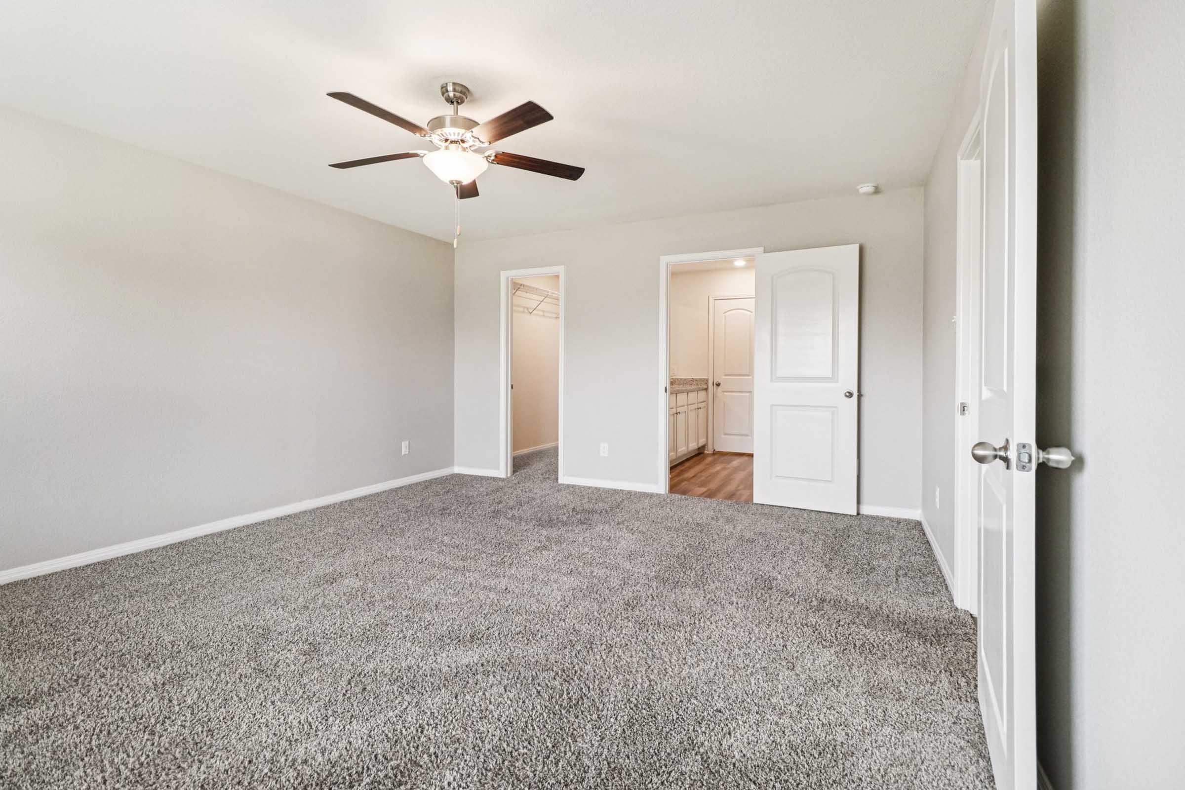 A spacious, empty bedroom featuring light gray walls, a ceiling fan, and plush carpet. There is a doorway leading to a closet on the left and another door on the right leading to a bathroom or additional room. Natural light fills the space, creating a bright and inviting atmosphere.