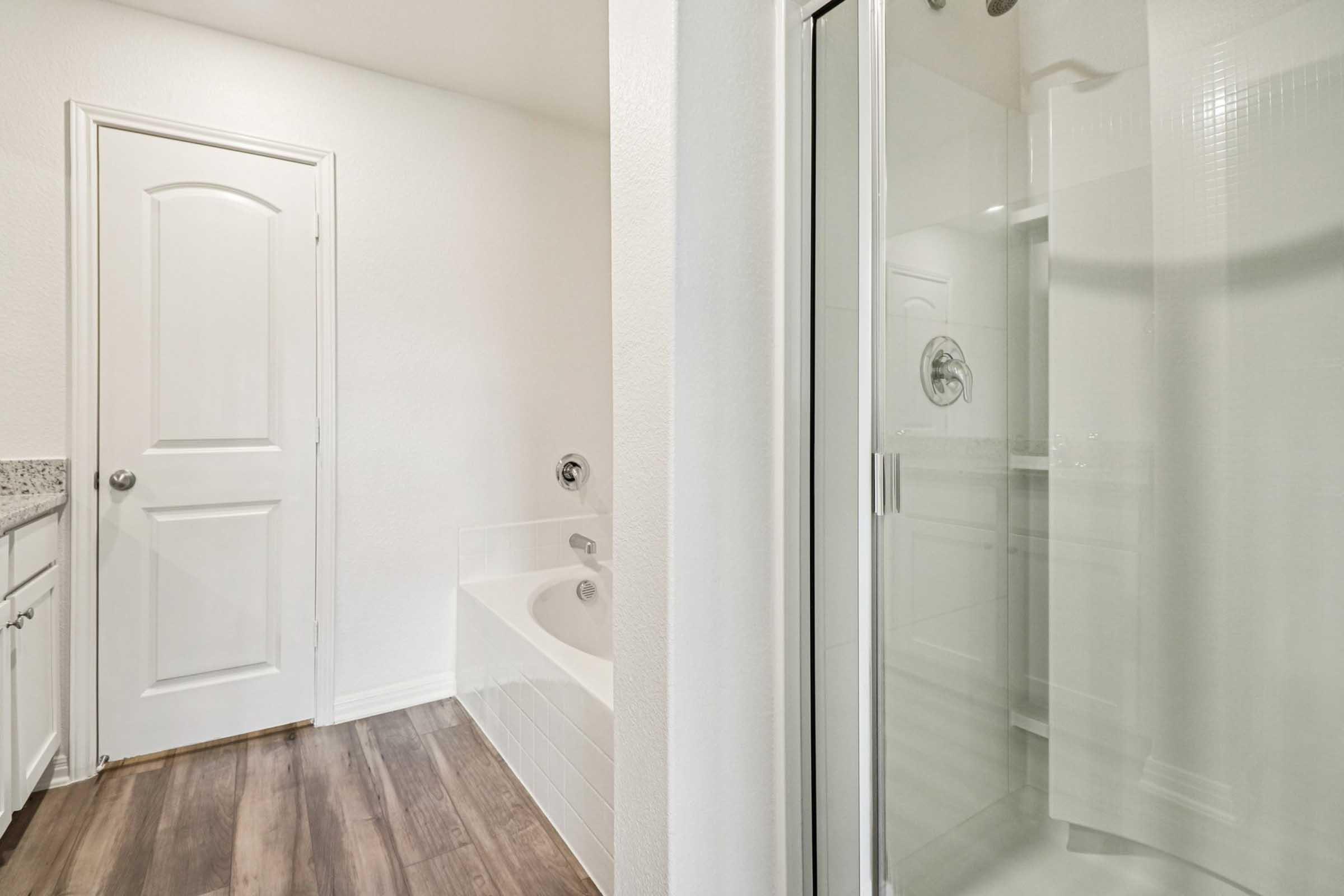 Interior view of a modern bathroom featuring a white door, a bathtub, and a glass shower enclosure. The walls are painted white, and the flooring is a wood-style laminate. The bathtub is adjacent to the shower, and a countertop with sink is visible in the background.