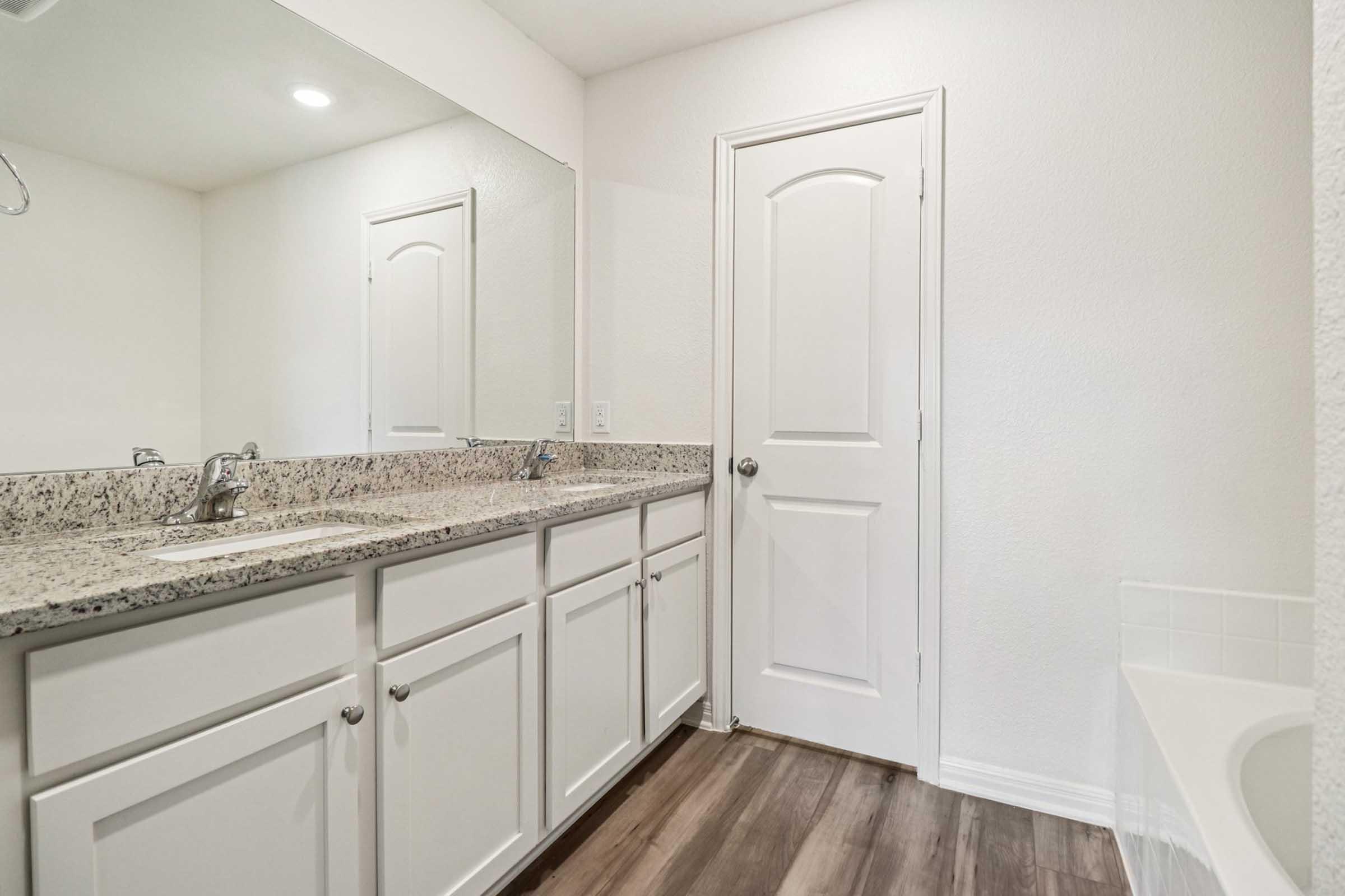 A spacious, modern bathroom featuring a double sink vanity with granite countertops, white cabinets, and a large mirror. The room has light-colored walls and flooring, with a bathtub located in the corner and a closed door leading to another area. Soft overhead lighting enhances the bright and clean atmosphere.