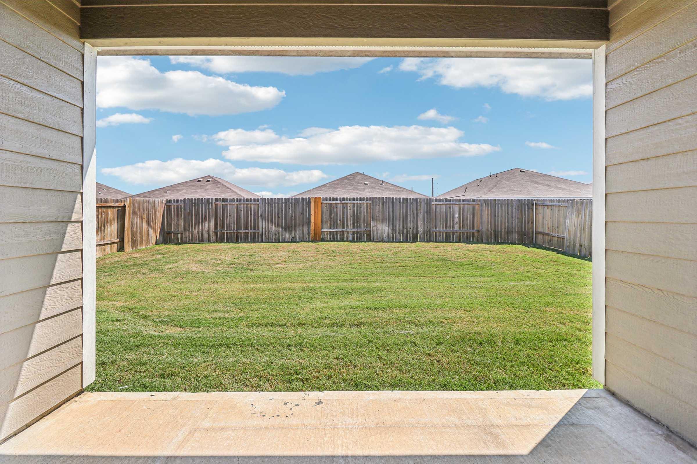 View from a covered patio of a backyard, featuring a neatly mowed lawn bordered by wooden fences. In the background, several houses with sloped roofs are visible against a bright blue sky with white clouds.