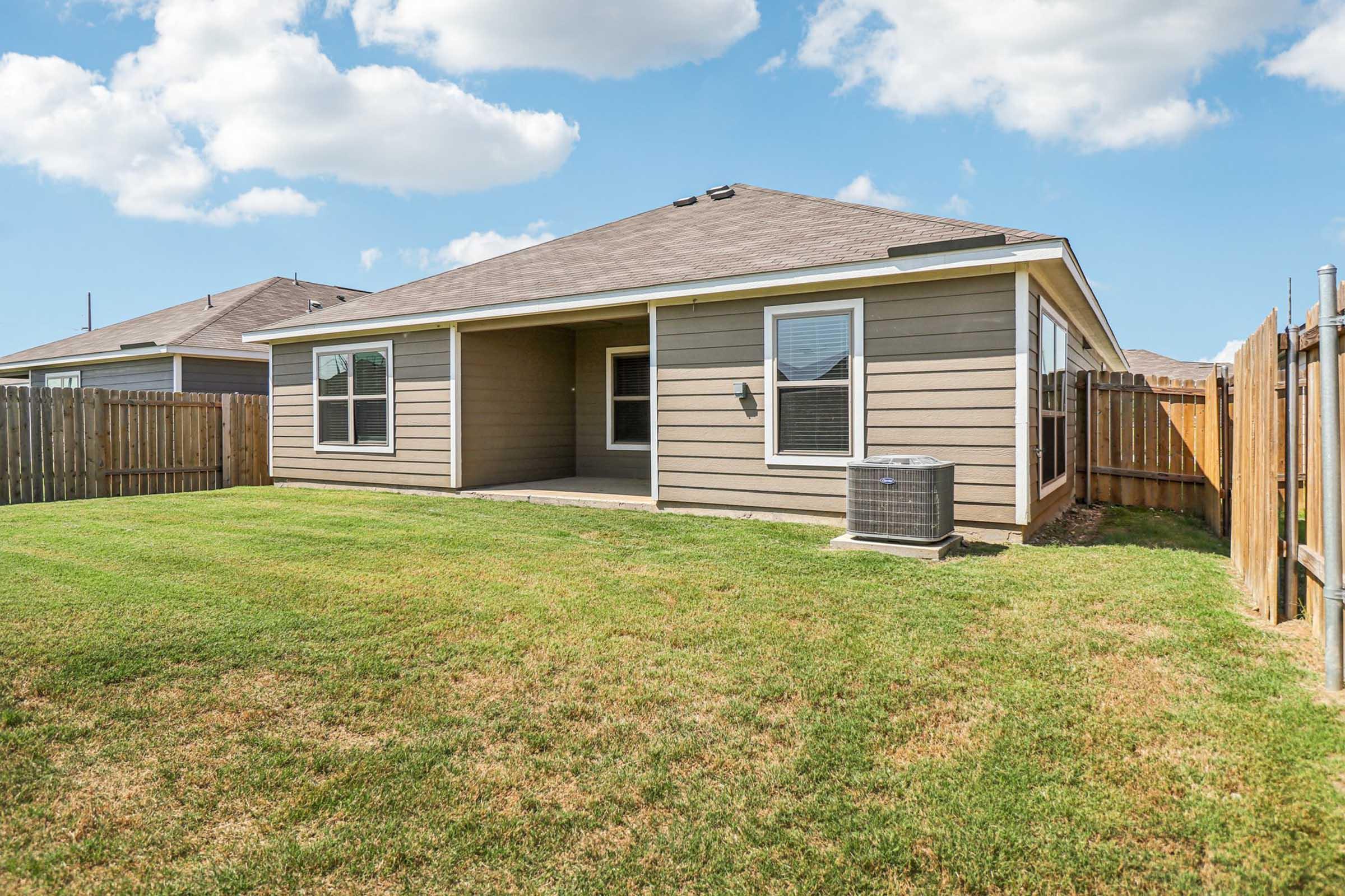 A well-maintained backyard featuring a grassy lawn, a patio area with no furniture, and a central air conditioning unit. The house has beige siding with large windows and is enclosed by a wooden fence, under a bright blue sky with fluffy white clouds.