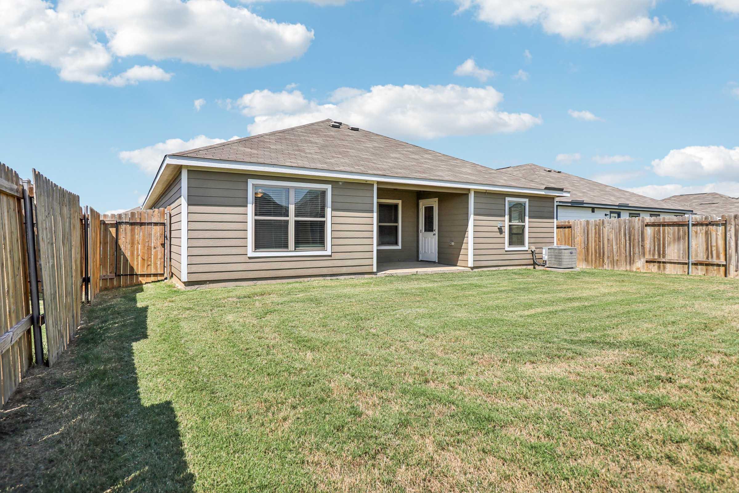 A single-story house with a light-colored exterior, featuring multiple windows, situated in a backyard with a neatly maintained lawn. The yard is enclosed by wooden fencing, and there's an air conditioning unit visible on the side. The sky is clear with a few clouds.