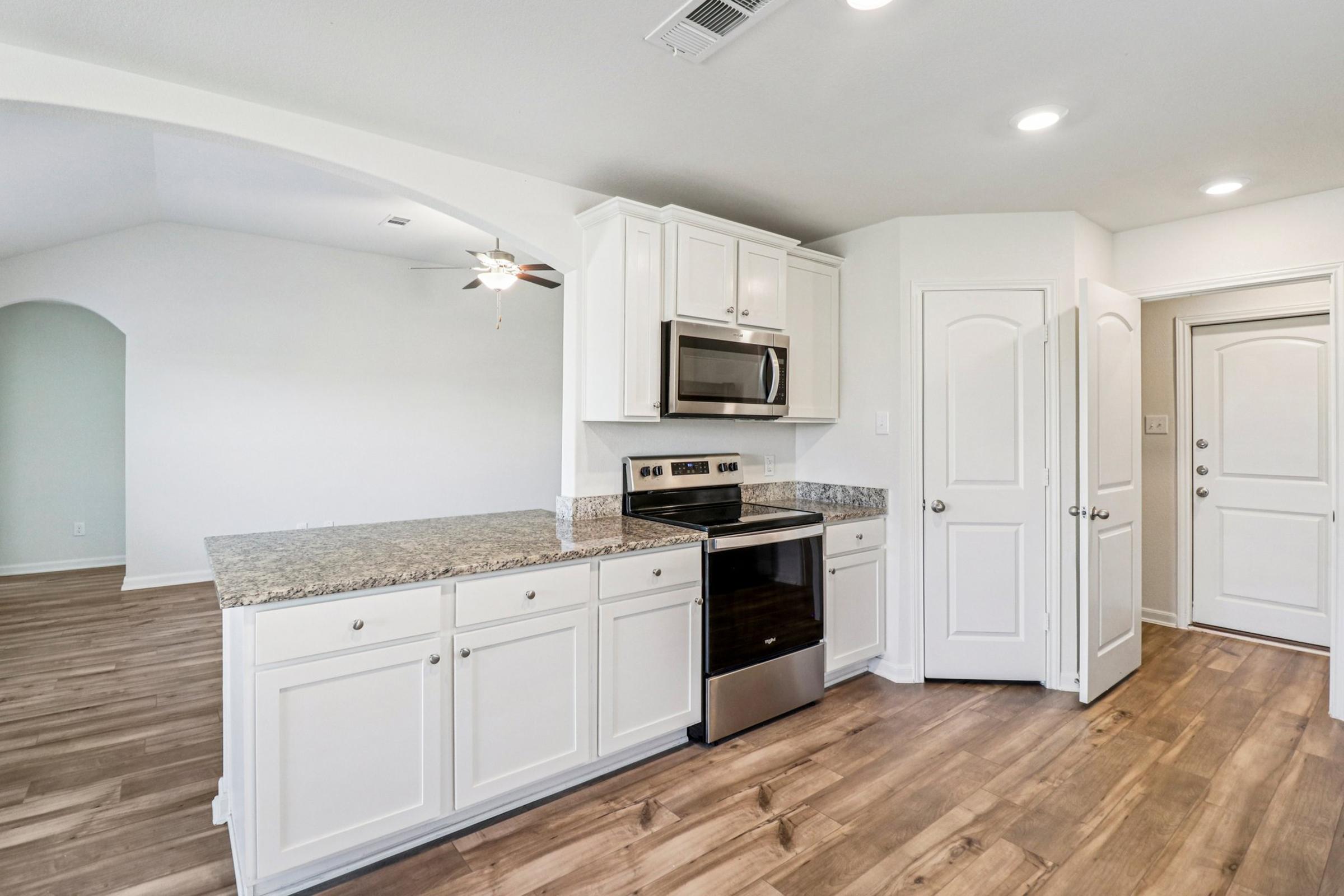 Modern kitchen featuring white cabinetry, stainless steel appliances, and granite countertops. Open layout with a view of a spacious living area, illuminated by natural light. Hardwood flooring throughout and a doorway leading to an entrance.