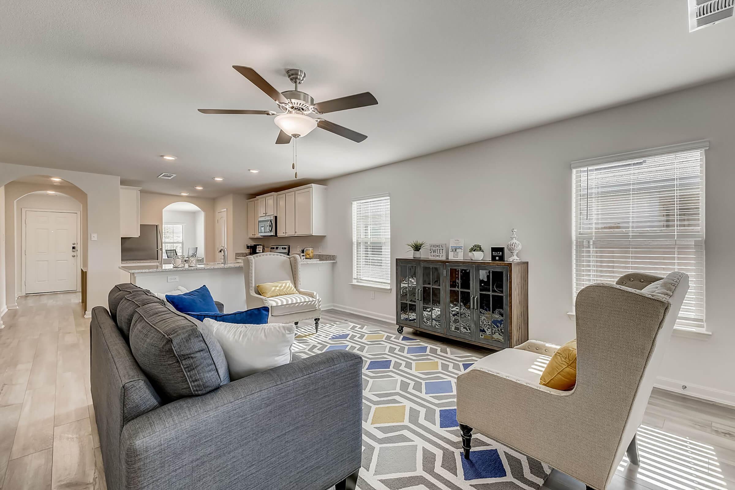 A modern living room featuring a gray sofa with colorful throw pillows, a stylish armchair, and a decorative cabinet. Natural light fills the space through large windows, highlighting the geometric patterned rug and the open layout connecting to the kitchen area.