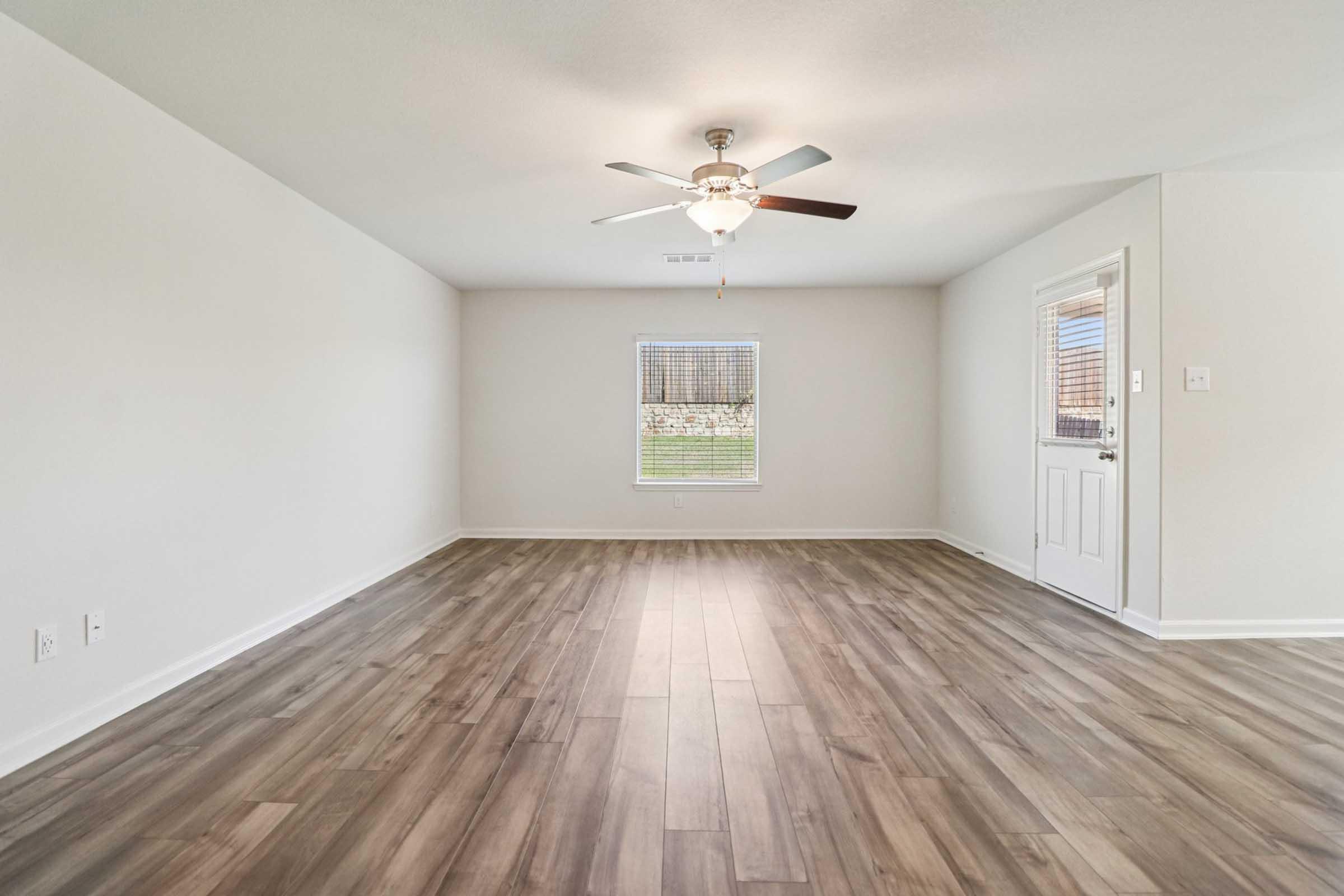 A bright, empty room with light-colored walls and wooden flooring. A ceiling fan is installed in the center, and there is a window on the left allowing natural light in. A door on the right leads outside, creating a welcoming and open space.