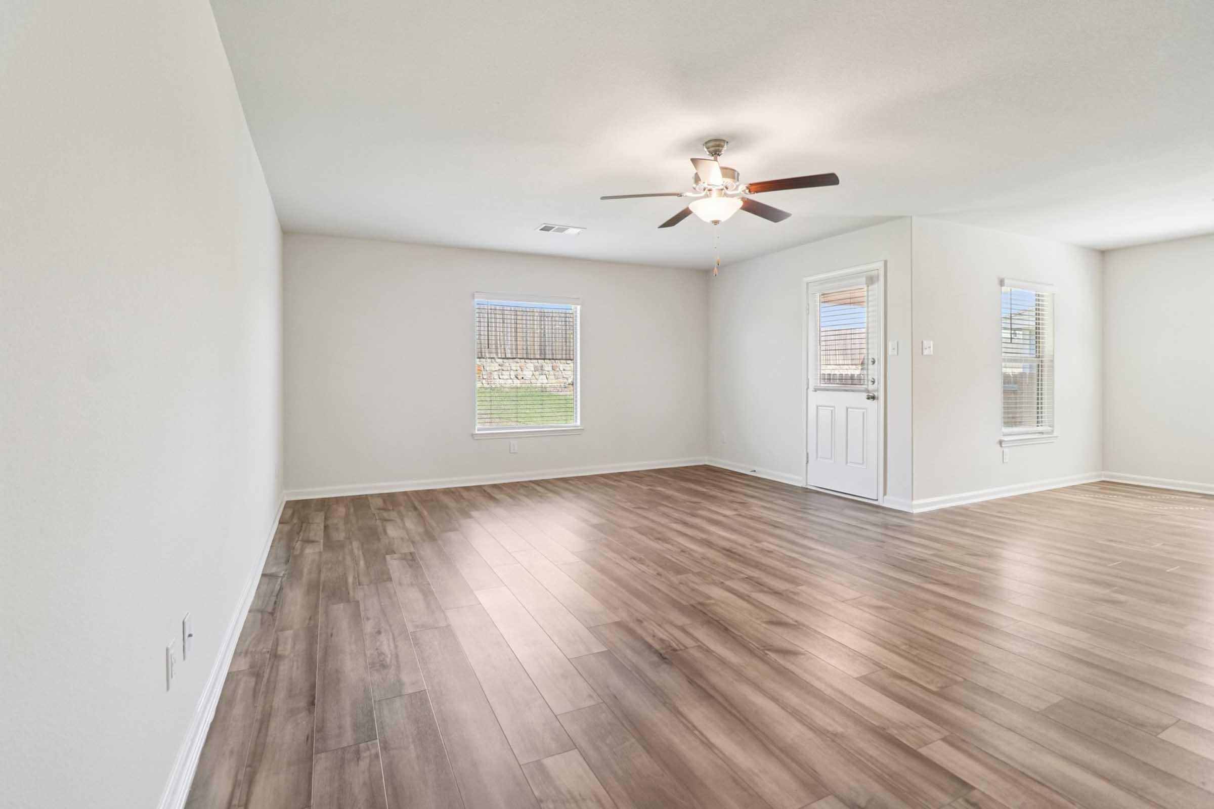 Empty room with light-colored walls and hardwood flooring. A ceiling fan is mounted overhead, and there is a door leading outside. Two windows allow natural light to enter, creating a bright, open space.