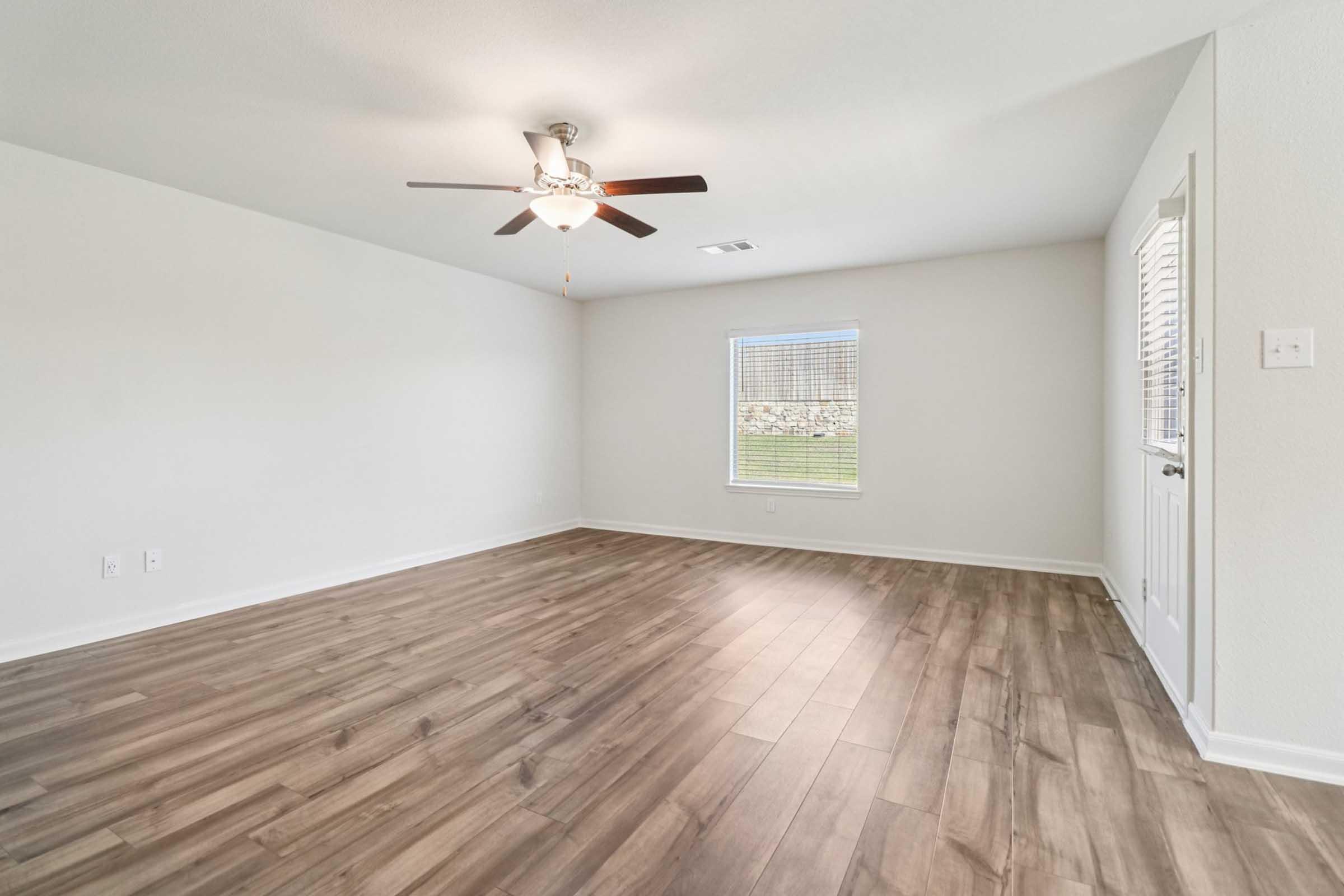 Spacious, vacant living room featuring hardwood-style flooring, a ceiling fan, and neutral walls. A window allows natural light, and a door leads outside. The room is empty, providing a blank canvas for decoration and furnishing.