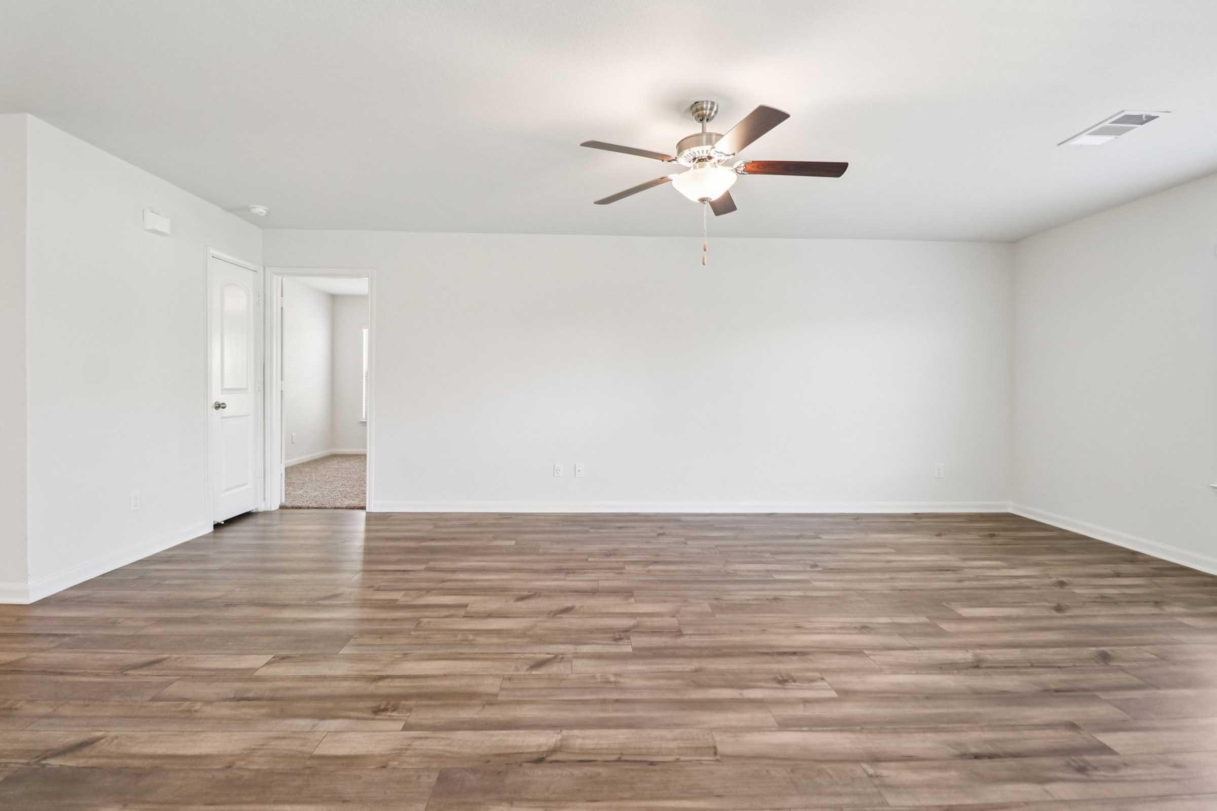 Spacious, empty living room featuring hardwood flooring, a ceiling fan, and a light-colored wall. A door on the left leads to another room, providing a view of the room's open layout and bright ambiance.