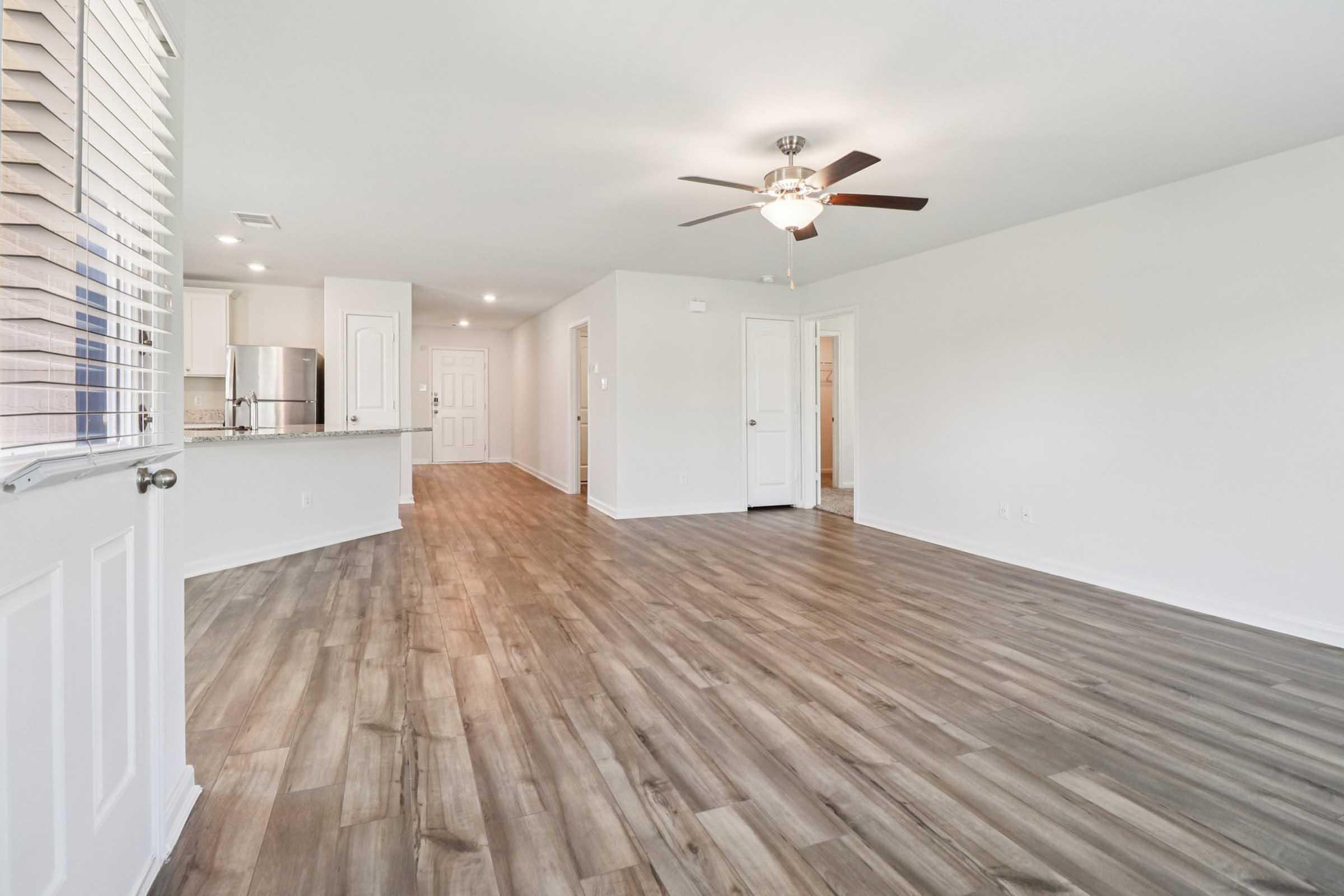 A spacious, modern living room featuring light-colored walls, a ceiling fan, and hardwood-style flooring. Natural light enters through the windows, and the open layout connects to a kitchen area in the background. The room is empty, emphasizing its roomy atmosphere.