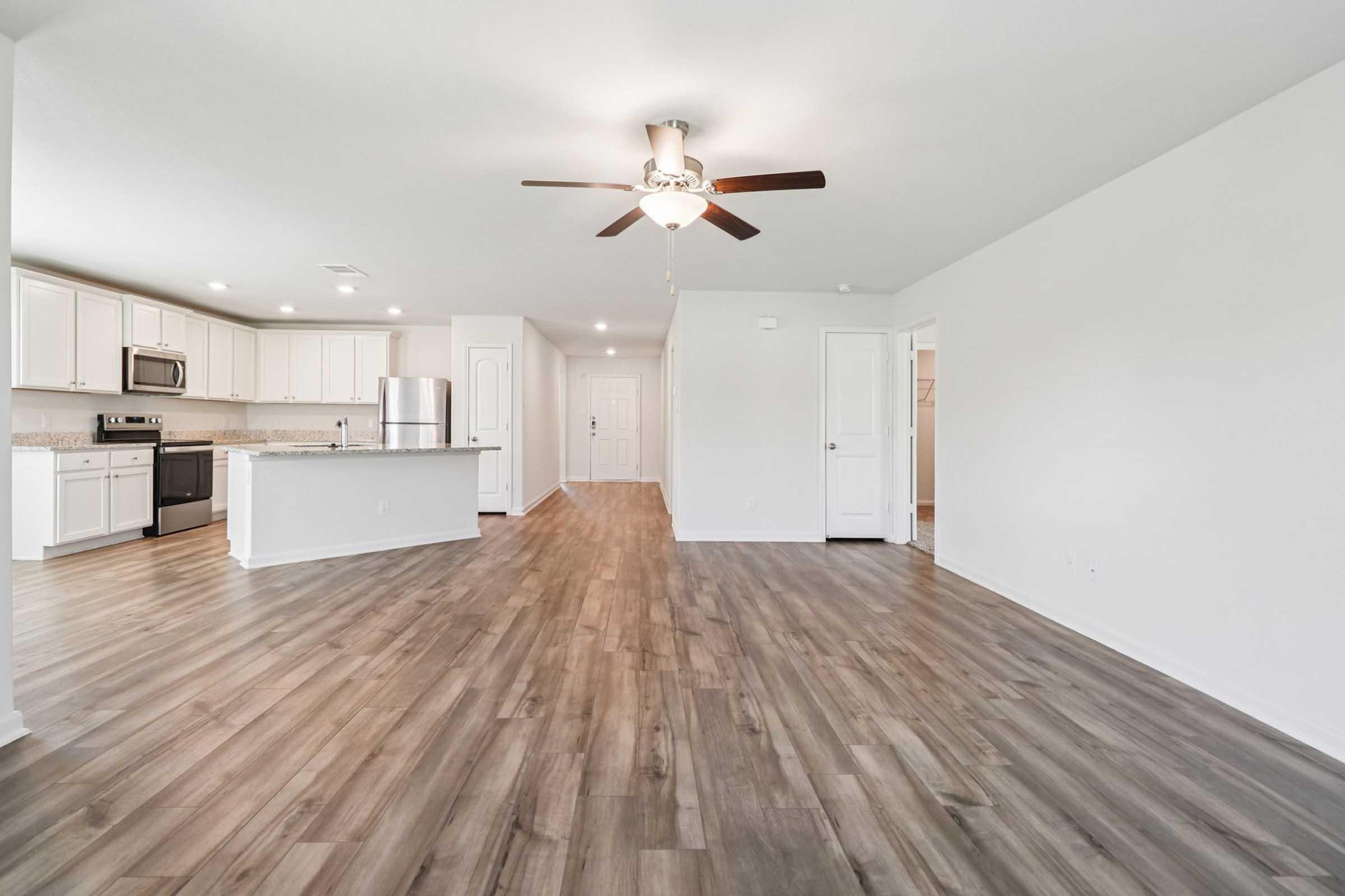 A spacious, modern living area with wooden flooring, featuring an open kitchen with white cabinets, stainless steel appliances, and a ceiling fan. The layout includes a door leading to a hallway and bright, neutral-colored walls, creating a welcoming atmosphere. Natural light enhances the cozy environment.
