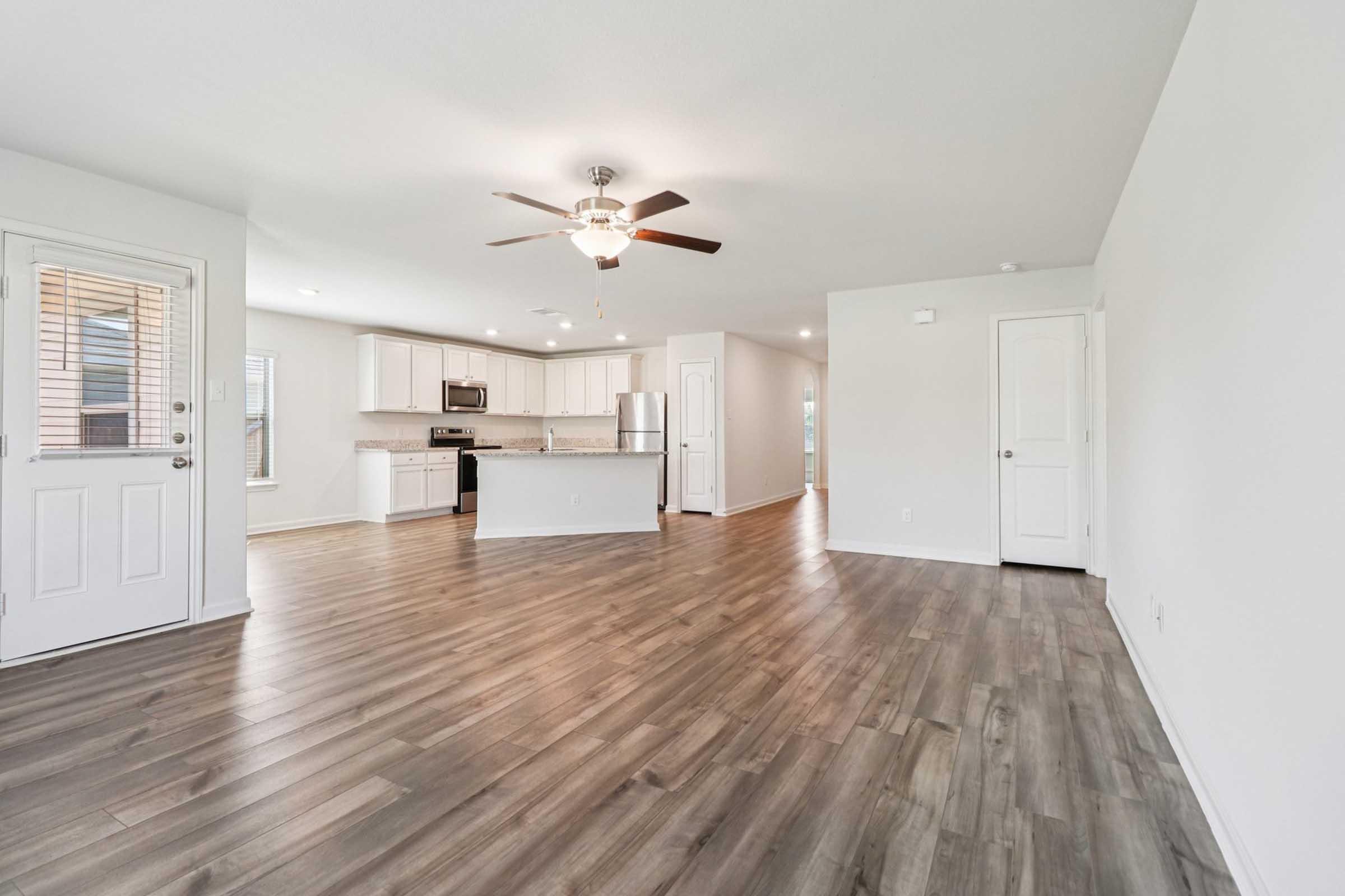 Spacious open-plan living area featuring light-colored walls and modern laminate flooring. The kitchen is equipped with stainless steel appliances and a central island. There's a ceiling fan, and large windows allow natural light. A front door leads outside, and another door opens to a hallway.