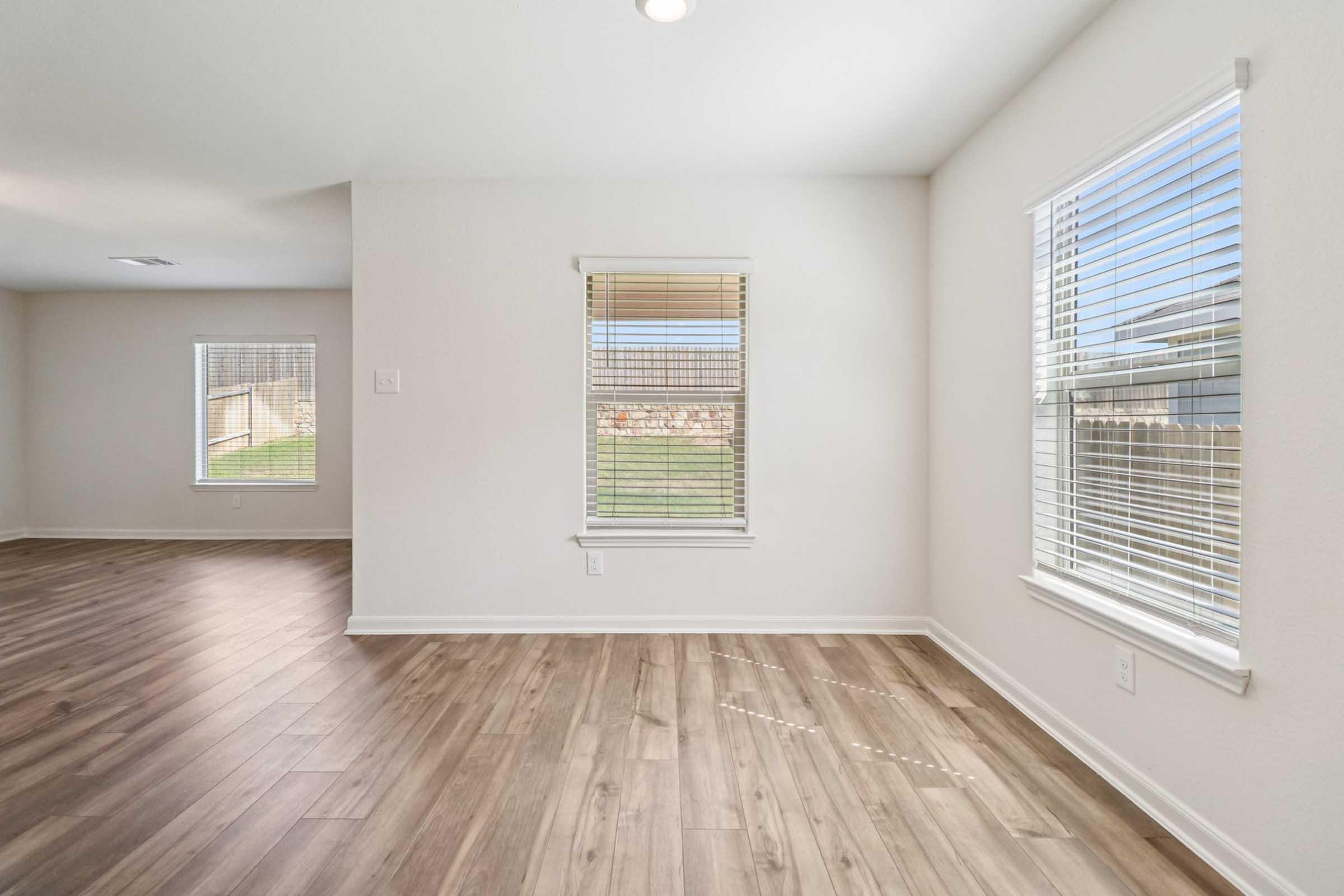 Bright and spacious interior of a room with light-colored walls and wood-style flooring. Two windows with blinds allow natural light to enter, revealing a view of a grassy outdoor area. The layout appears open and inviting, showcasing modern design elements.